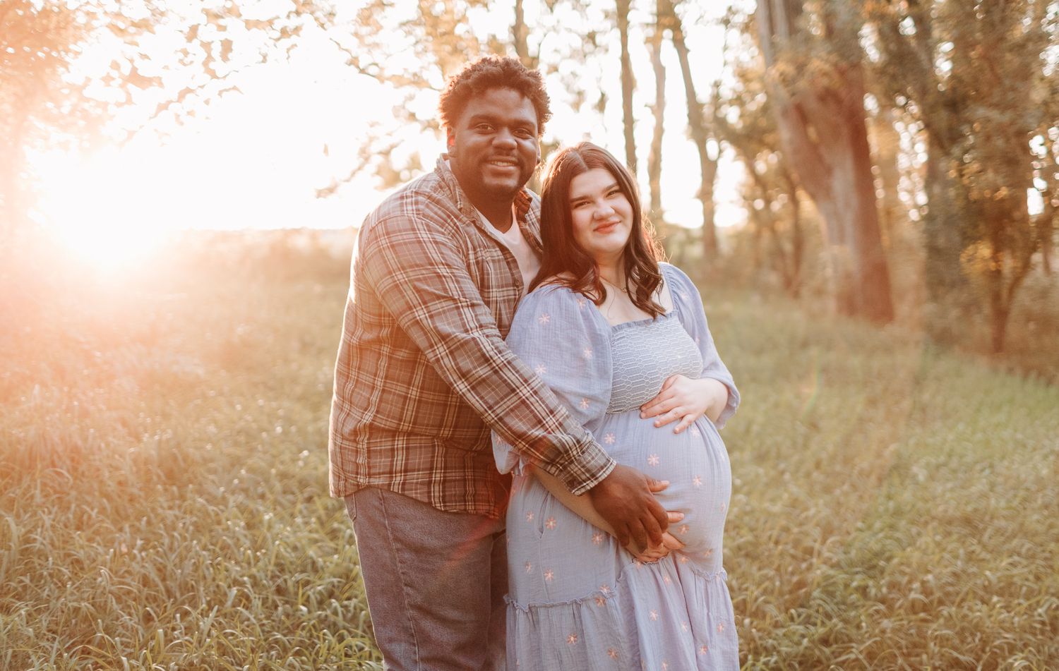 Couple maternity photo at golden hour in a field, woman in blue dress cradling baby bump.