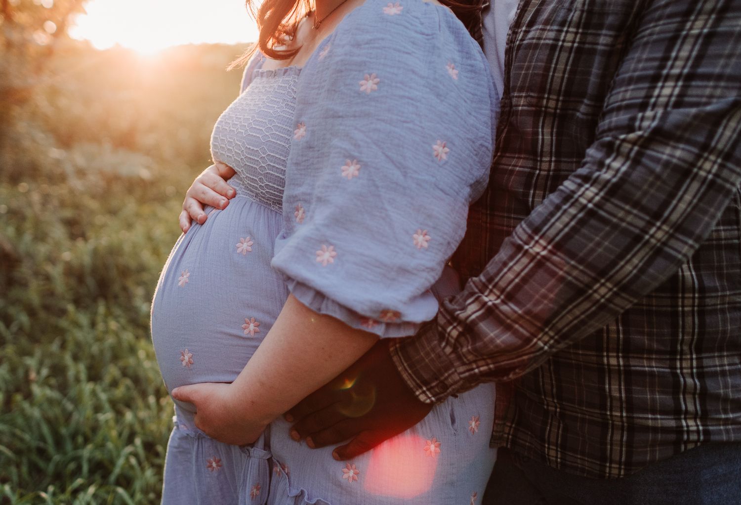 Close-up of pregnant woman in blue floral dress with partner embracing her at golden sunset outdoors.