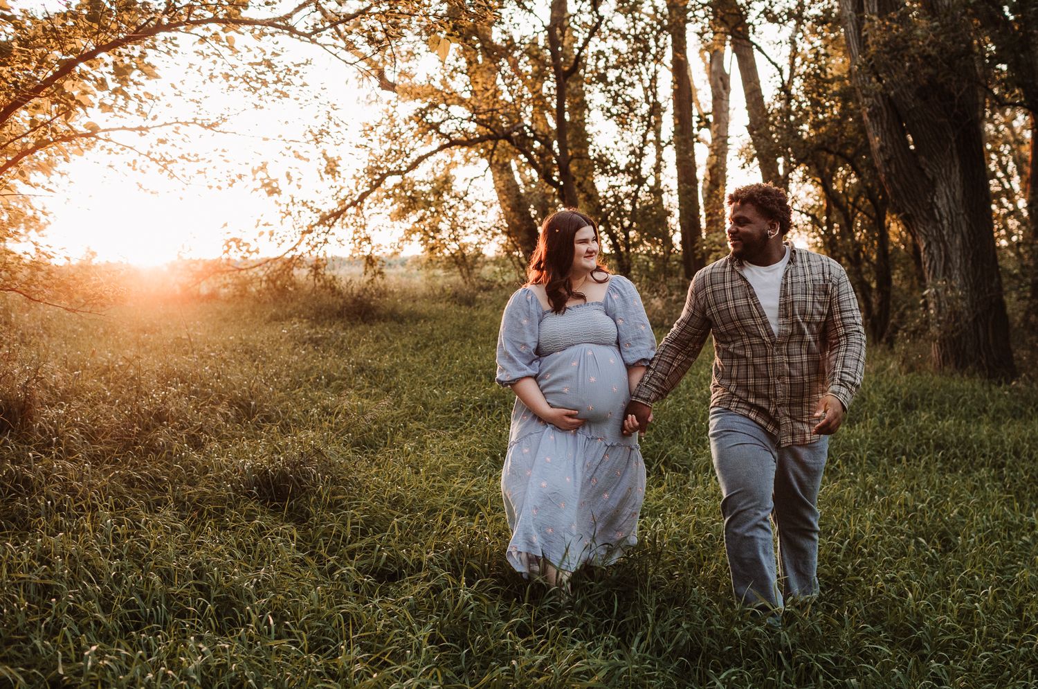 Pregnant couple walking hand-in-hand through a sunlit meadow at golden hour, woman in light blue dress.