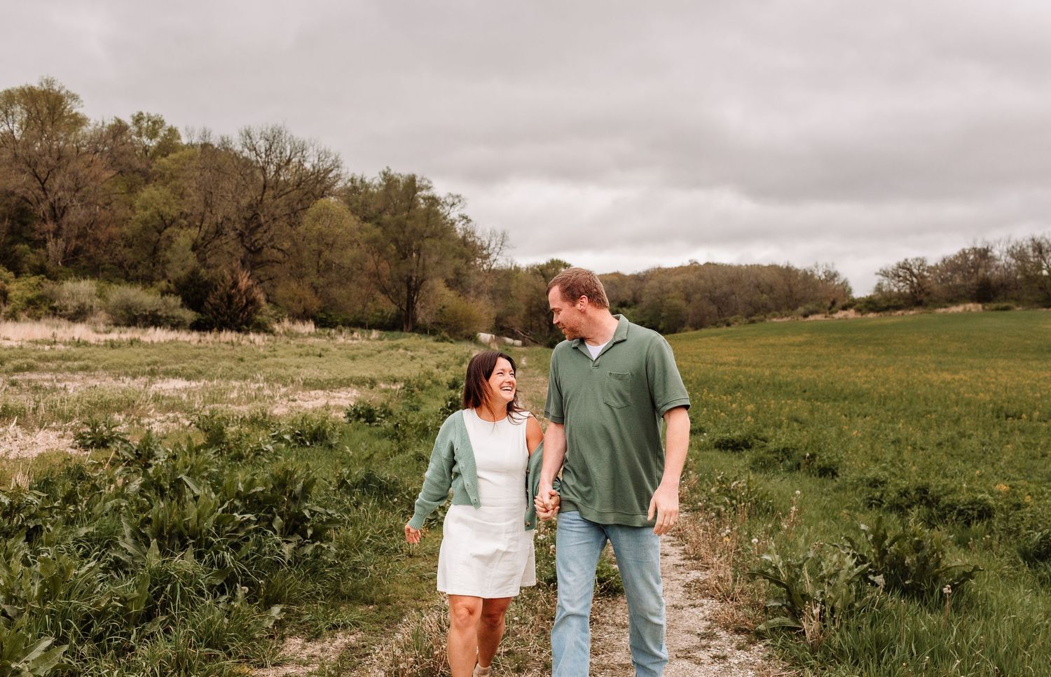 A couple holds hands walking on a dirt path through green fields under a cloudy sky.