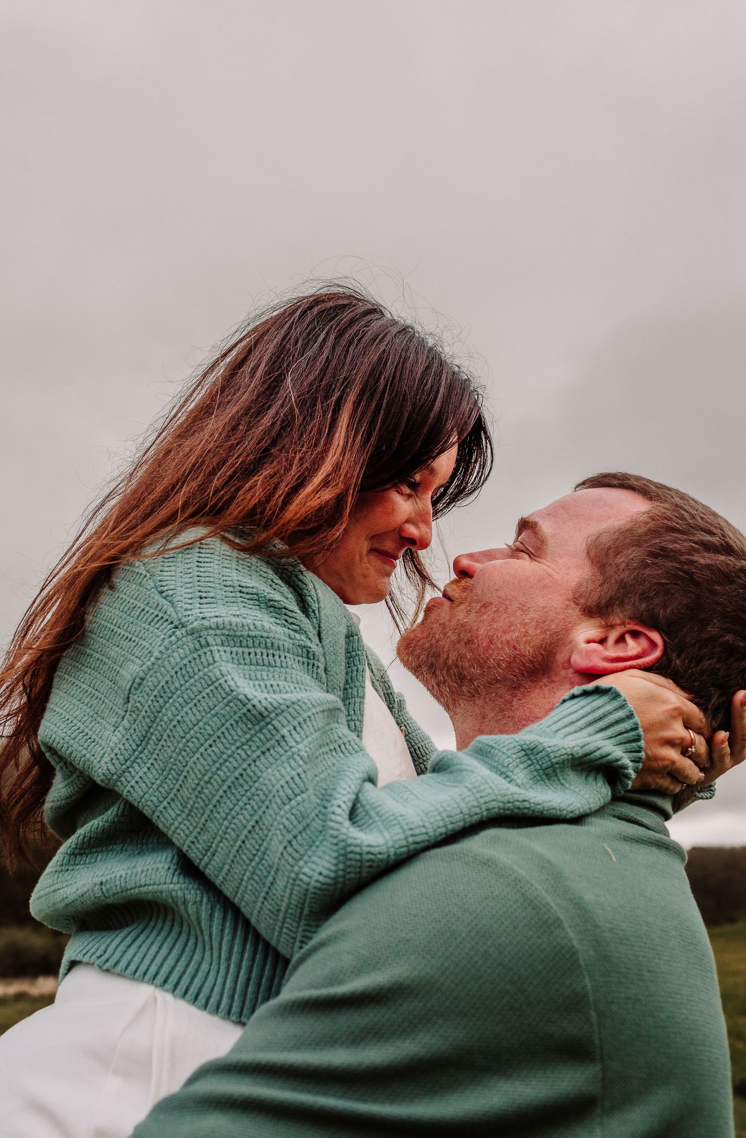 A couple in matching green sweaters share a romantic moment, gazing at each other outdoors on a cloudy day.