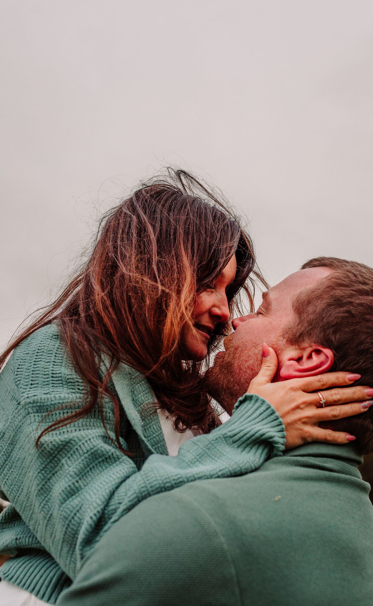 Romantic couple sharing a kiss outdoors, woman in teal cardigan leaning in close to man in green sweater.