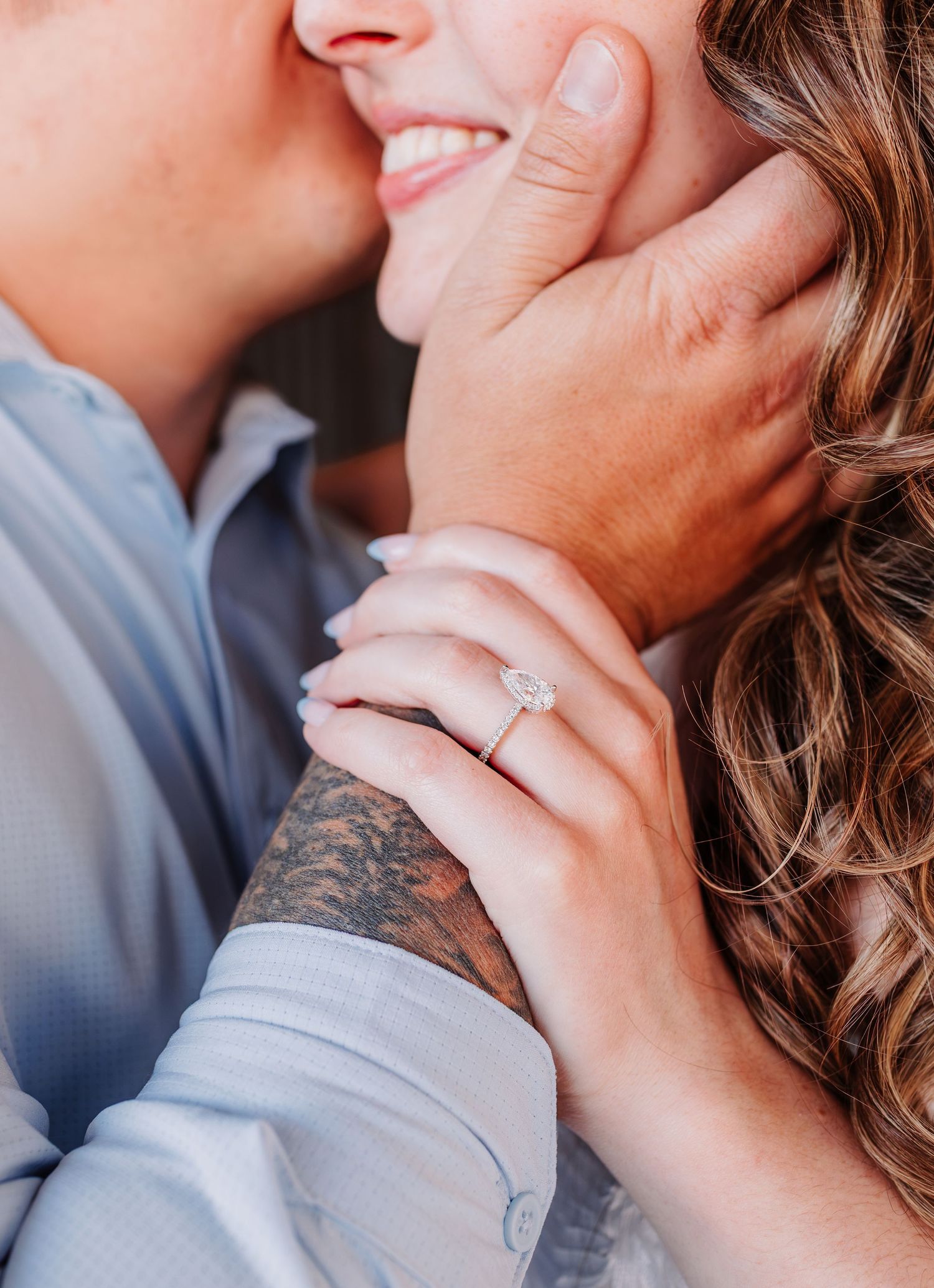 Close-up of smiling woman with diamond engagement ring on finger, embraced by tattooed partner in blue shirt.