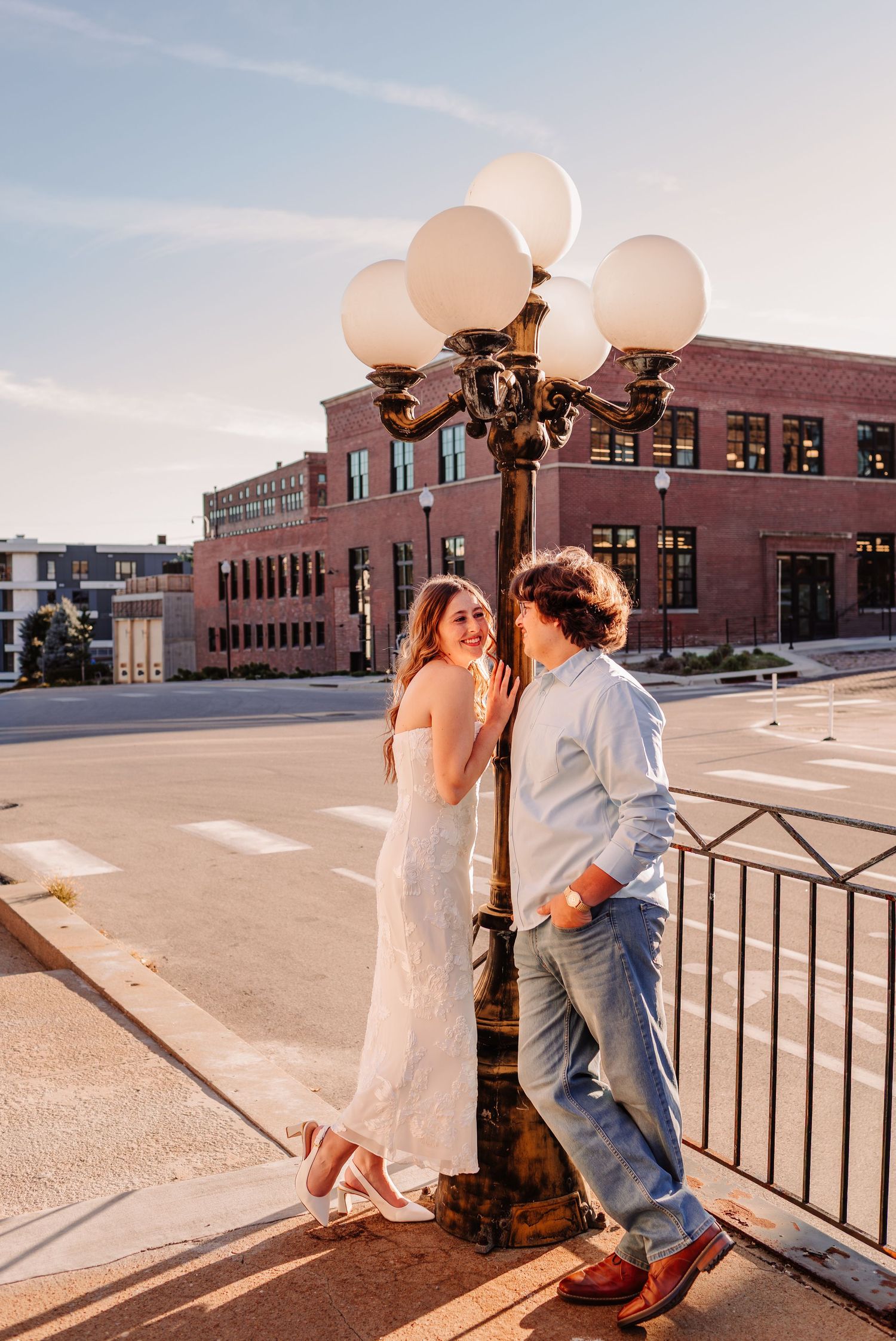Couple in white dress and jeans leaning against ornate street lamp at golden hour on urban bridge.