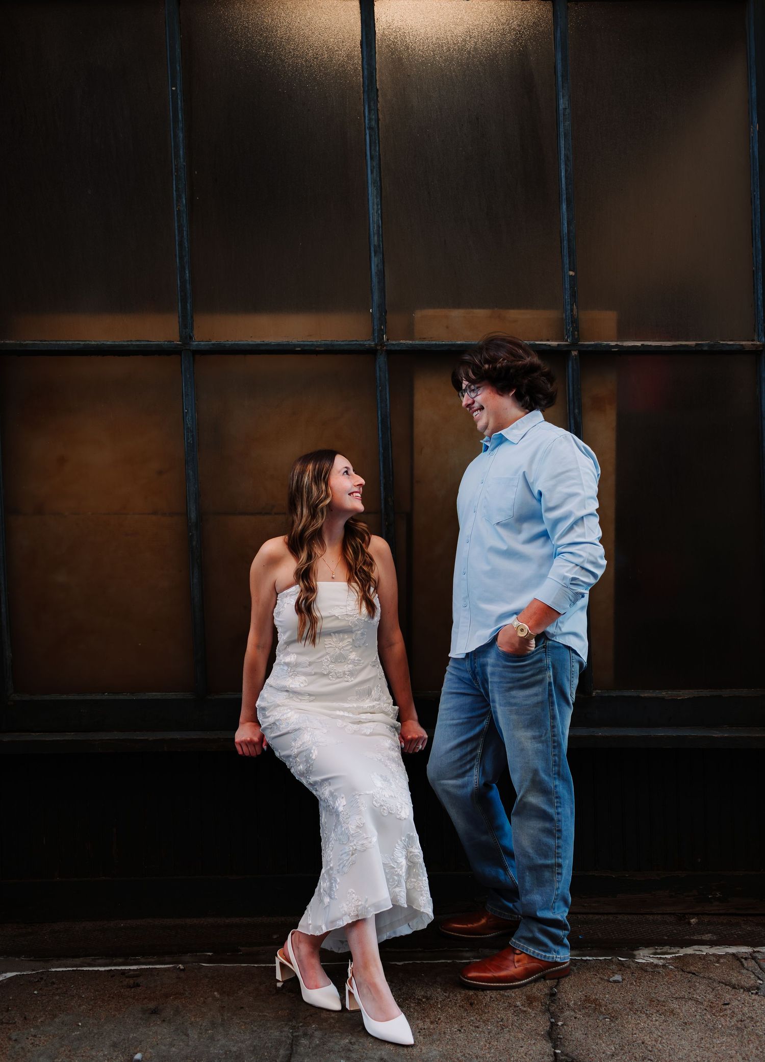 Couple holding hands and smiling at each other, woman in white dress and man in blue shirt, against dark industrial wall.