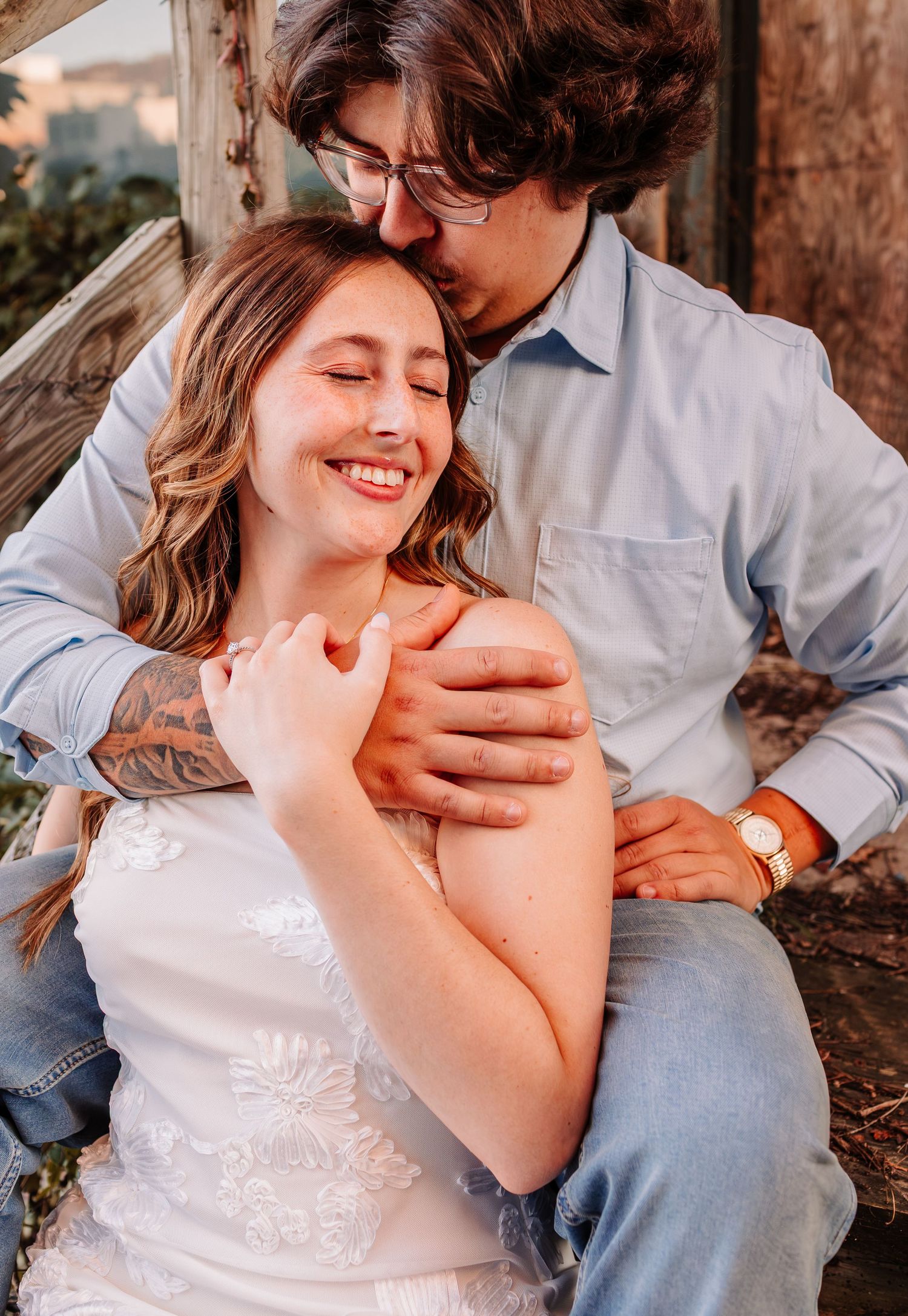 A couple shares a tender moment outdoors, with a man kissing a smiling woman on the head while embracing her shoulders.