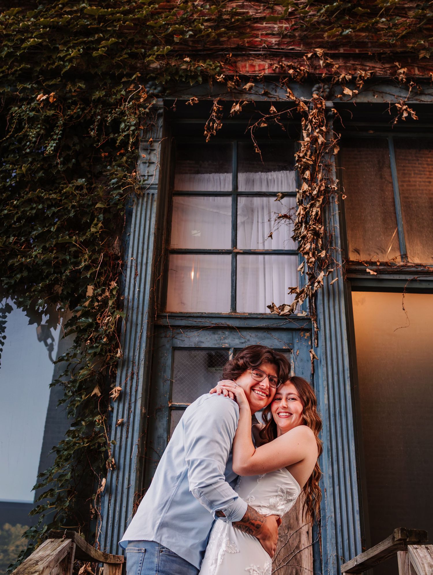 A couple embraces and smiles in front of a rustic ivy-covered building with large windows at golden hour.