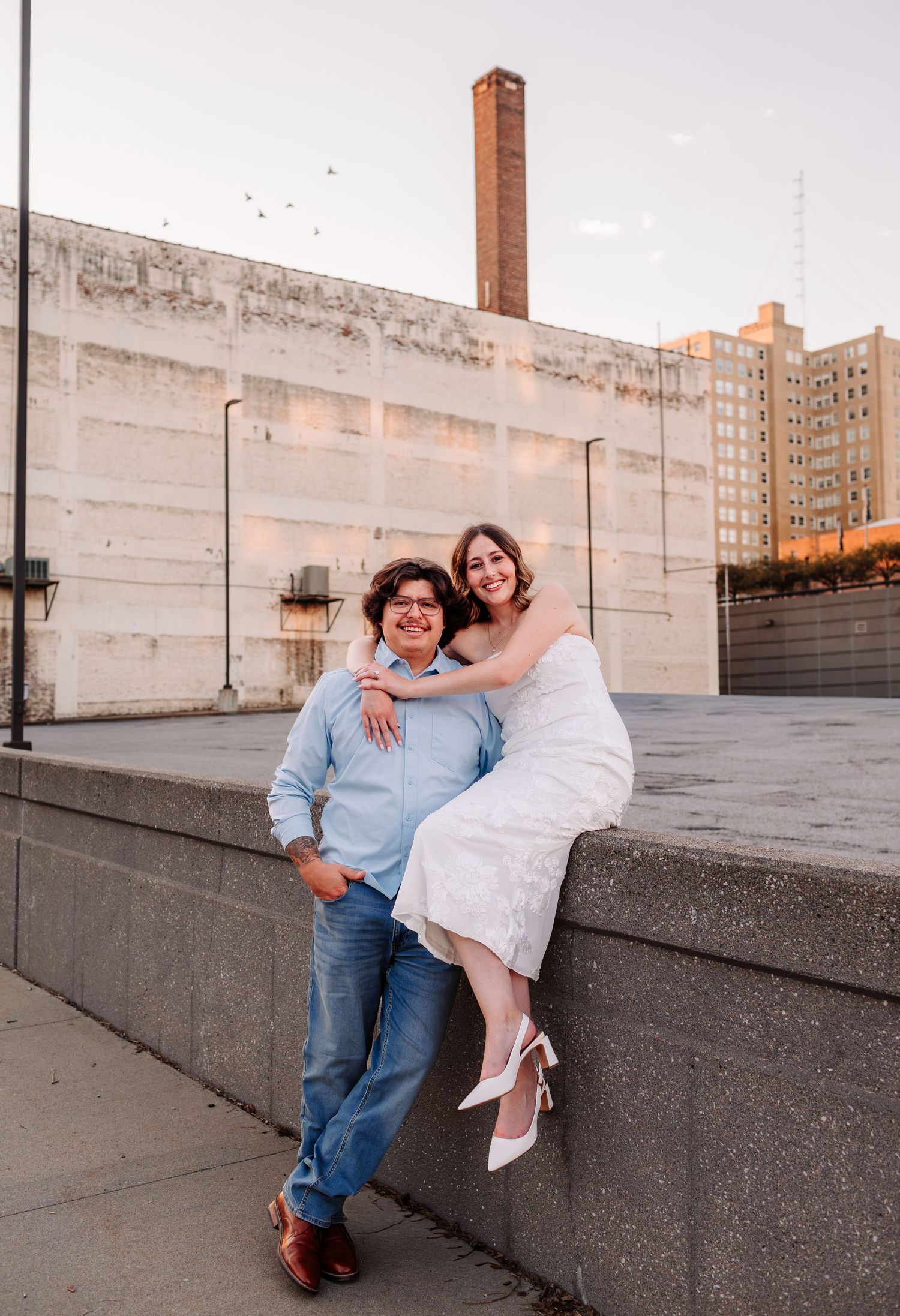 Couple posing on urban rooftop at sunset, woman in white dress sitting on ledge, man in blue shirt and jeans smiling.