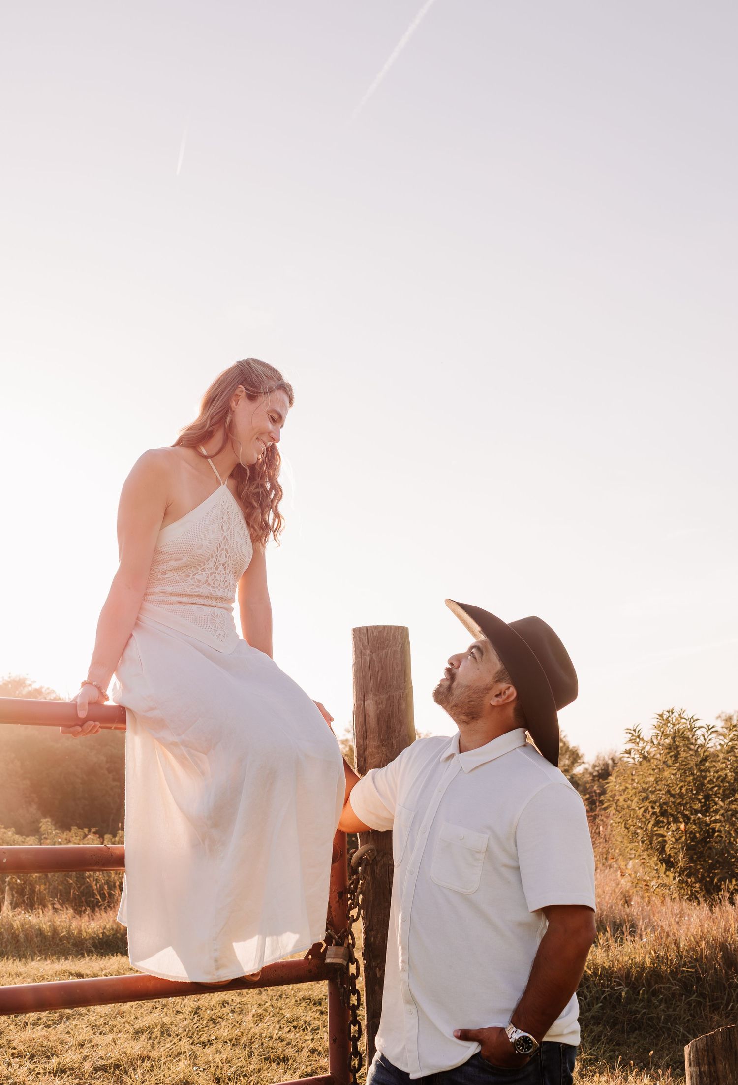 A woman in a white dress sits on a rustic fence while a man in a cowboy hat looks up at her during golden hour.