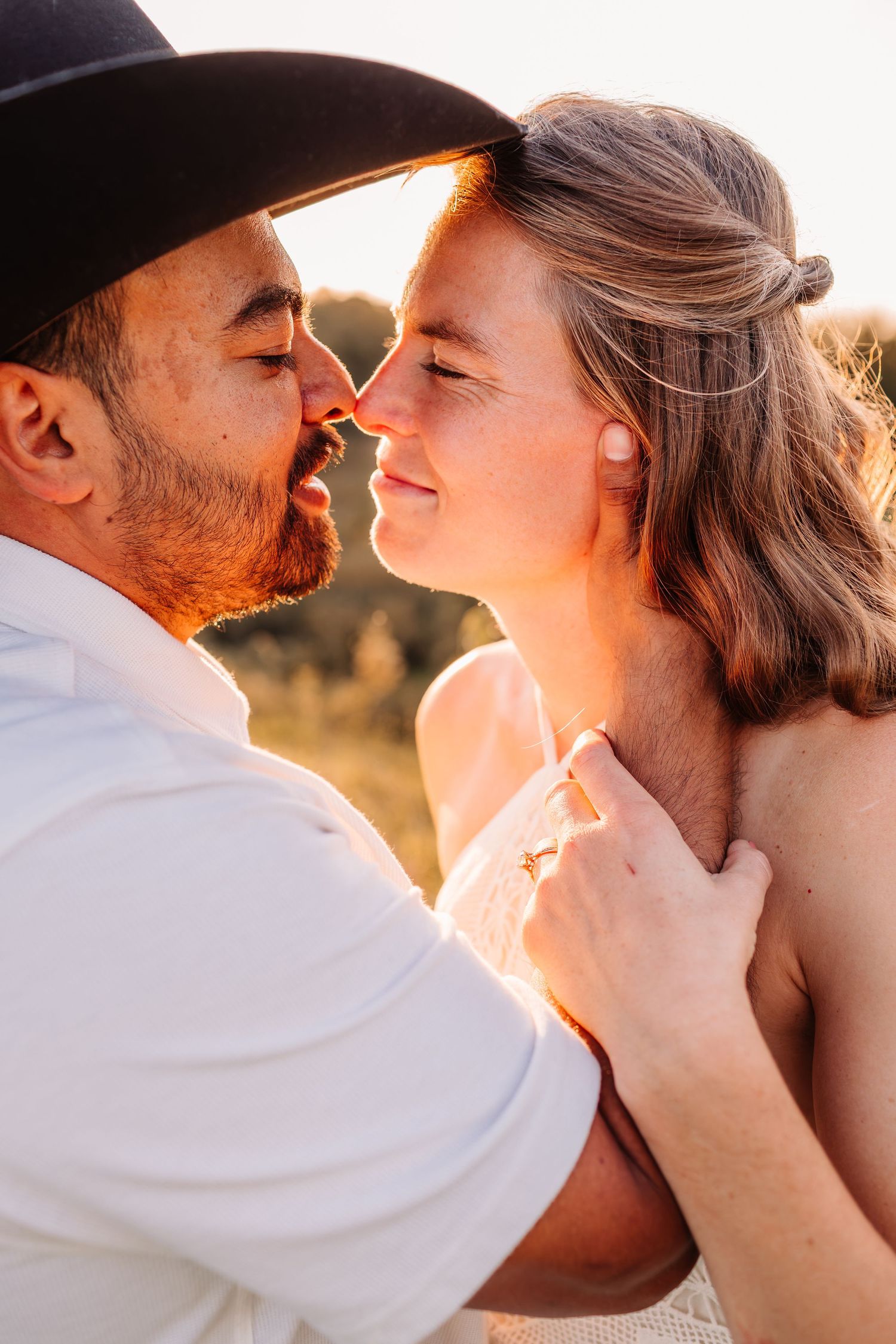 A couple shares a romantic almost-kiss at golden hour, the man wearing a black cowboy hat and white shirt outdoors.