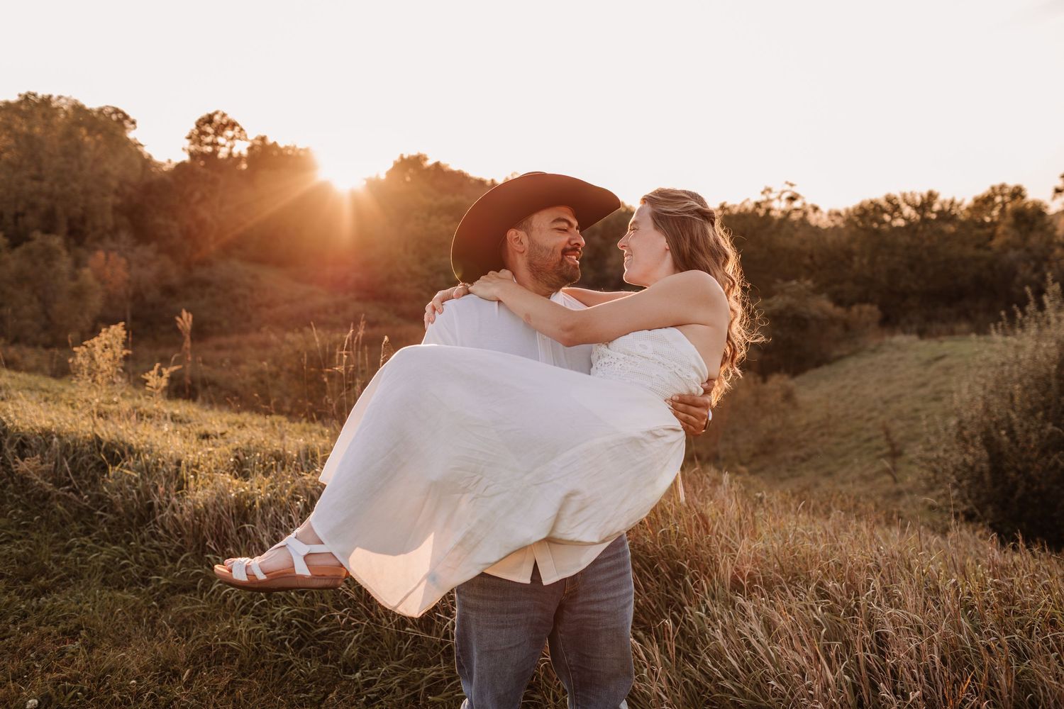 Groom in cowboy hat carries bride in white dress at golden sunset in a scenic field.