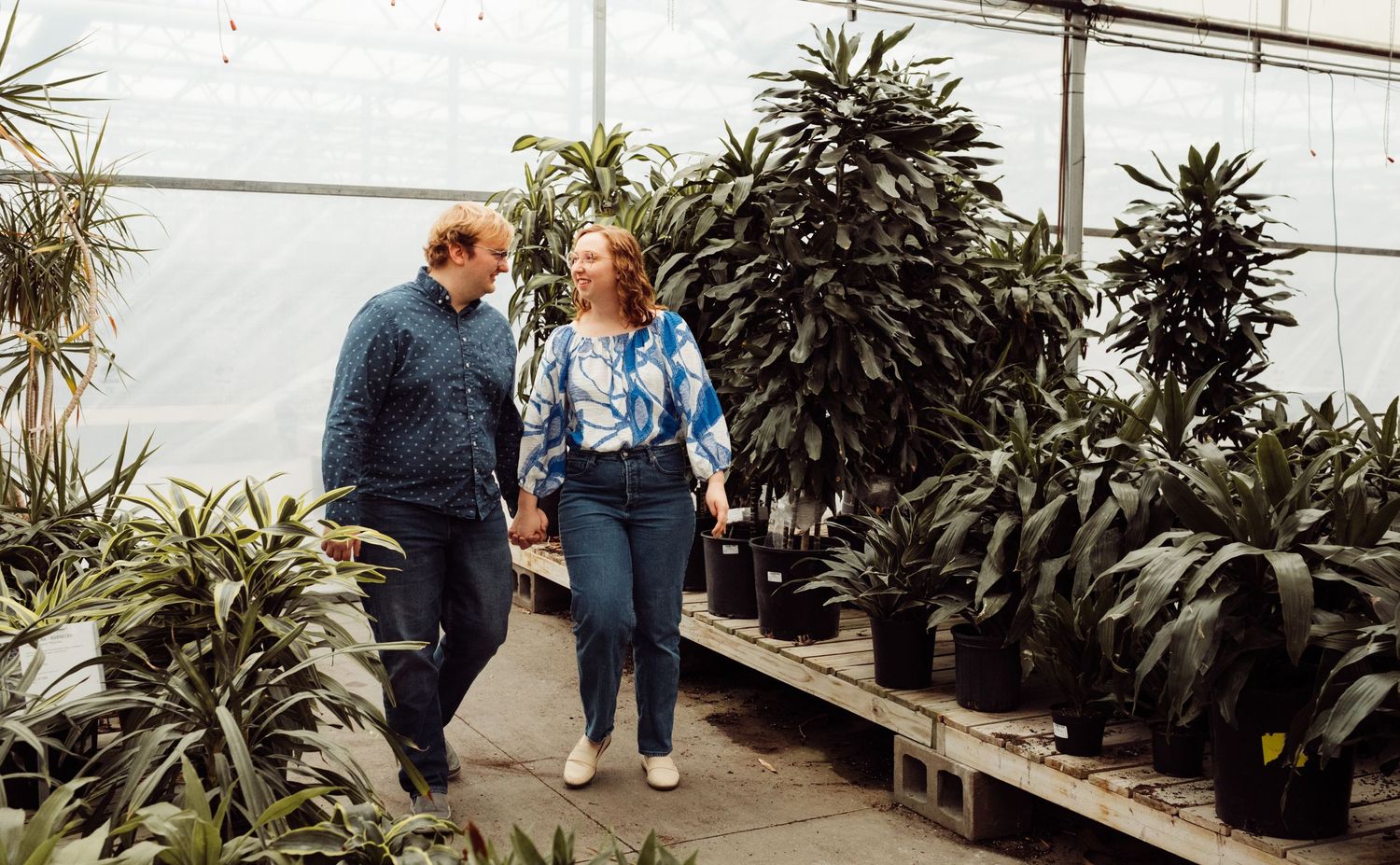 Couple walking hand in hand through a lush greenhouse filled with tropical green plants.