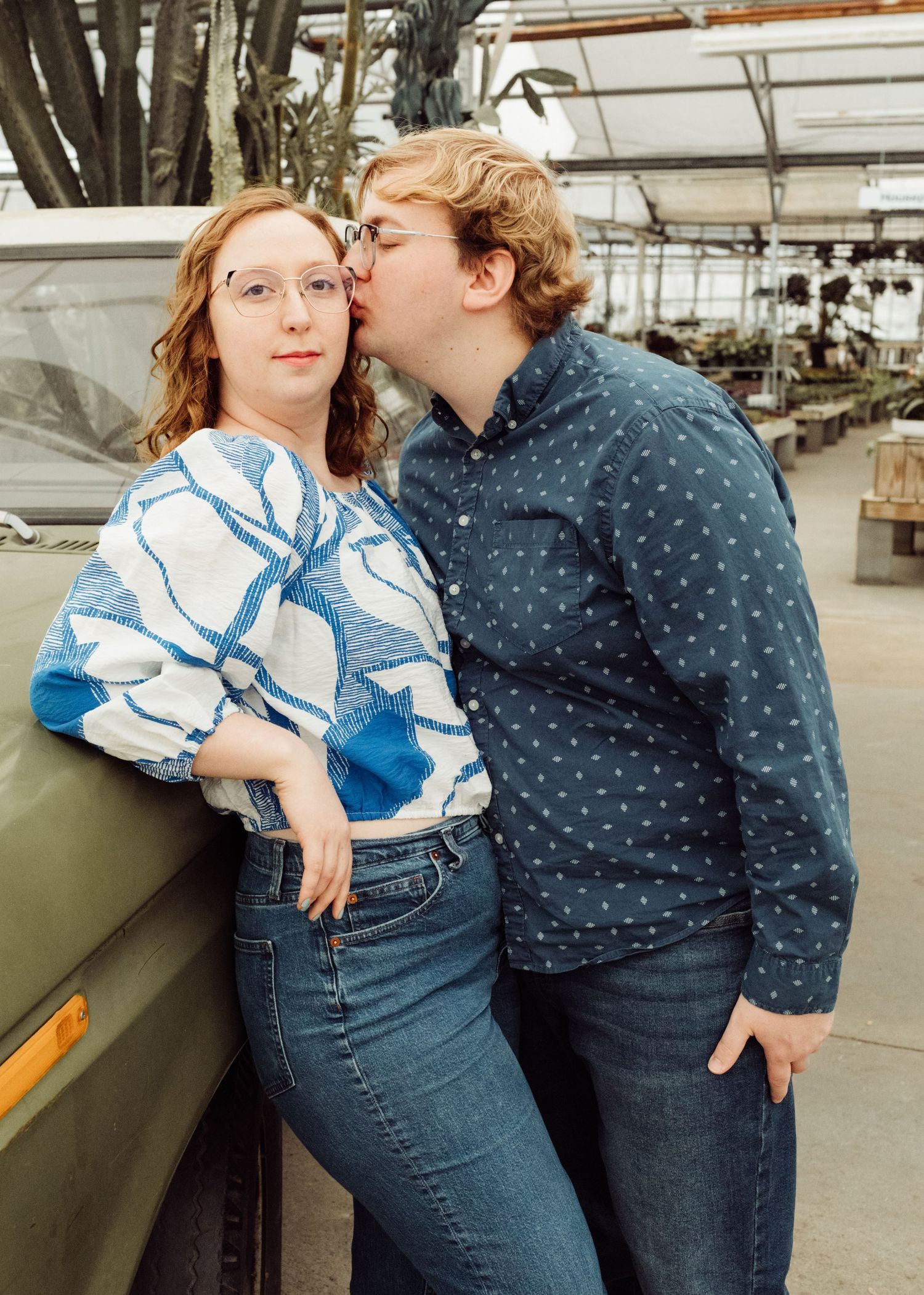 Young couple posing near a vintage green truck in a greenhouse, man kissing woman on the cheek.