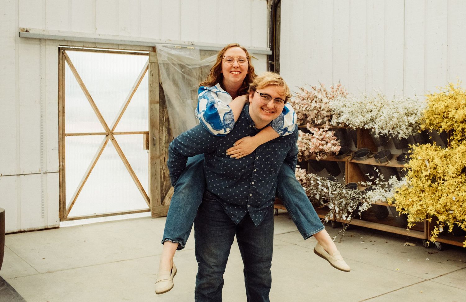 Man giving woman a piggyback ride in front of a white barn with colorful flowers in background.