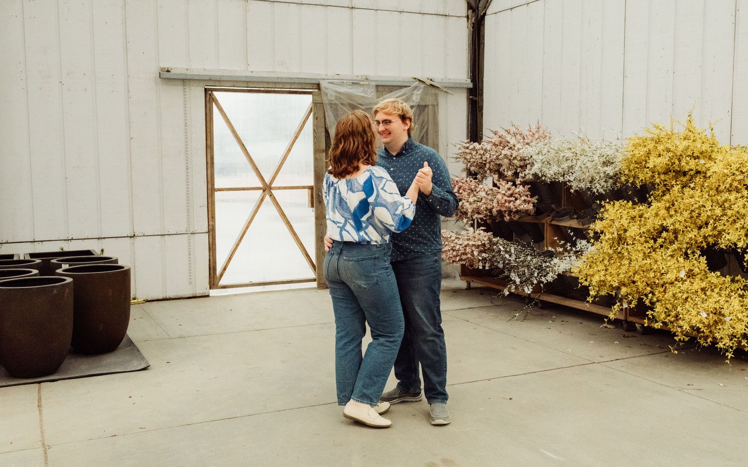 Couple dancing together outdoors in front of a white barn with colorful flowers and large pots nearby.