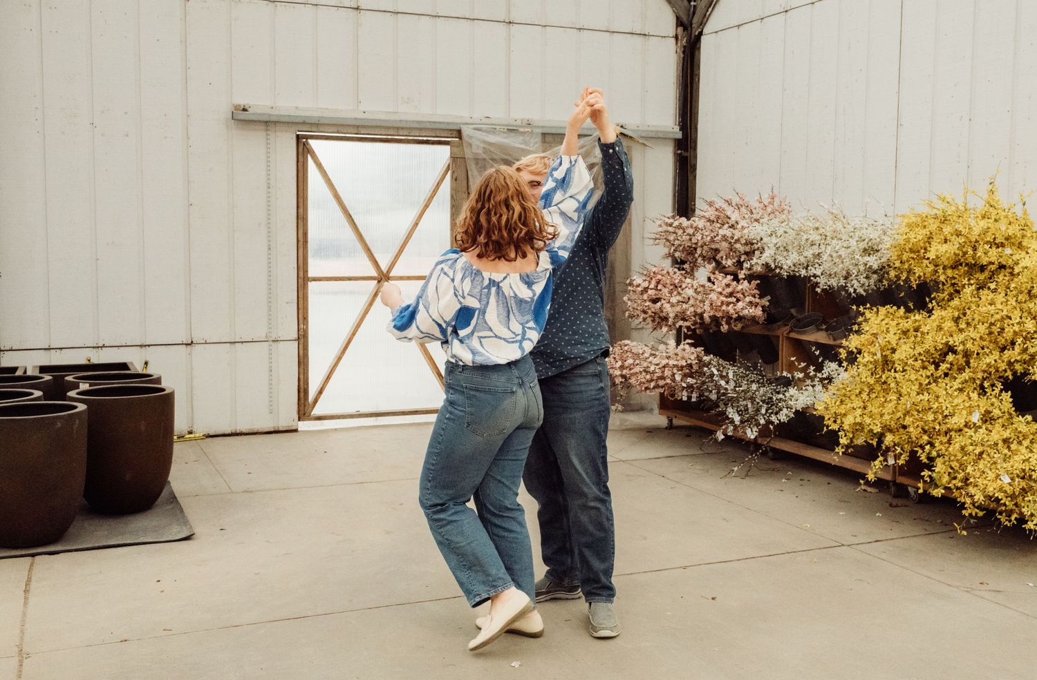 Couple spinning and dancing outdoors near a white barn surrounded by colorful blooming flowers.
