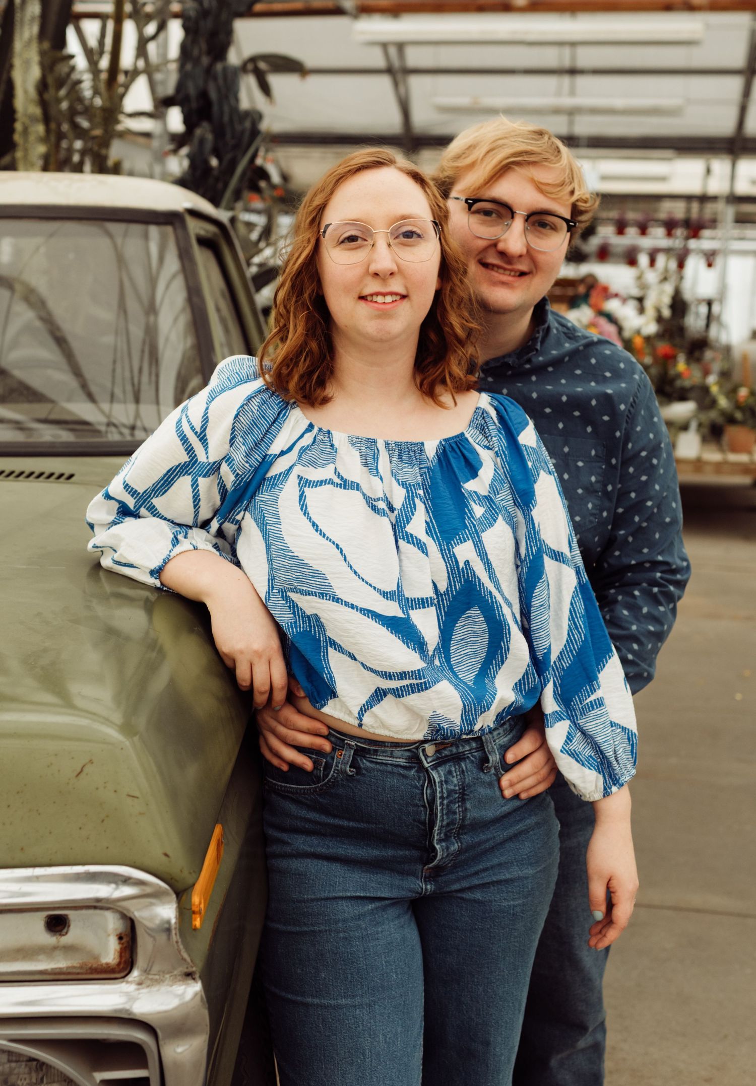 Smiling couple leaning against a vintage green truck inside a greenhouse with flowers behind them.
