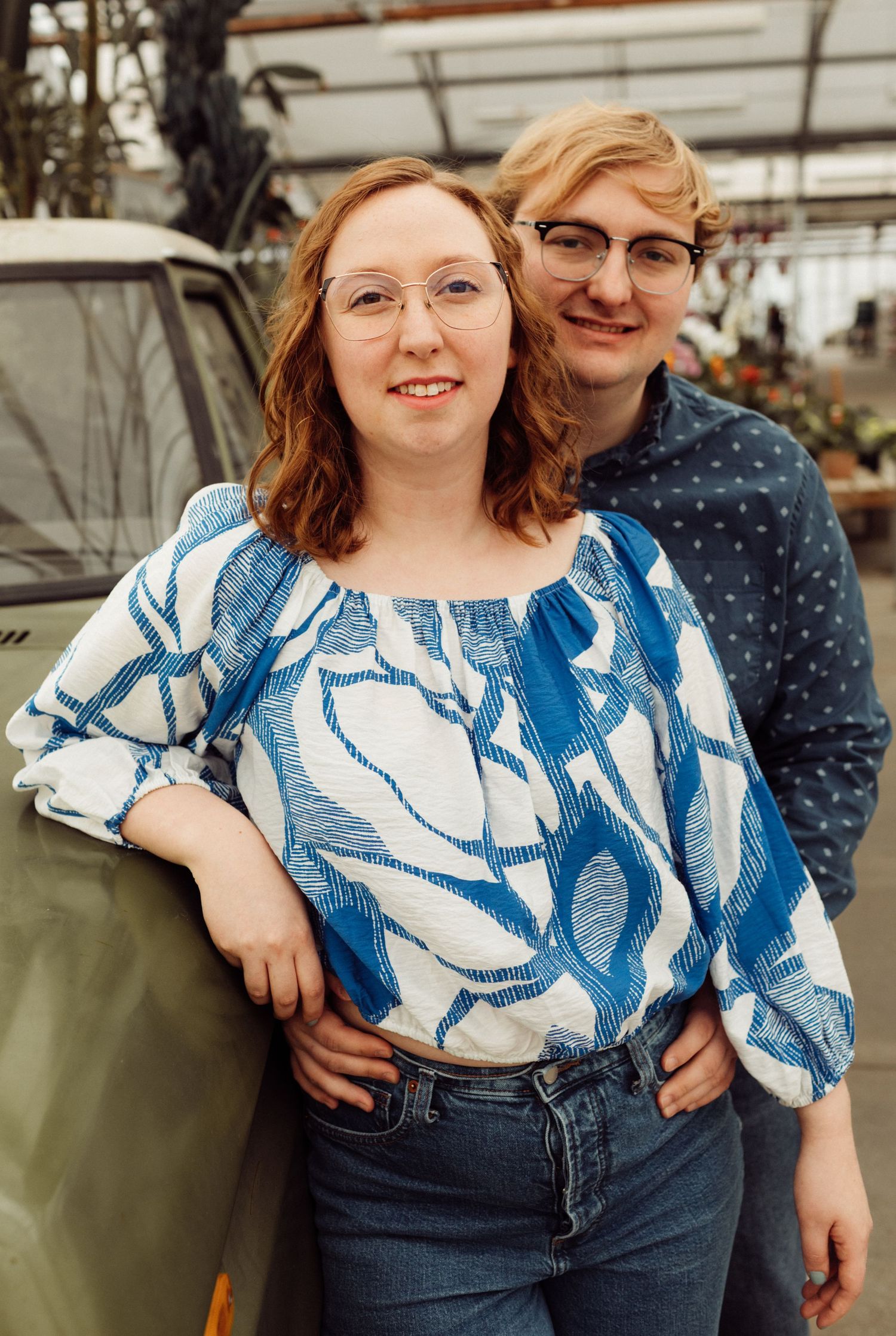 Young couple posing together in greenhouse near vintage green vehicle surrounded by plants.