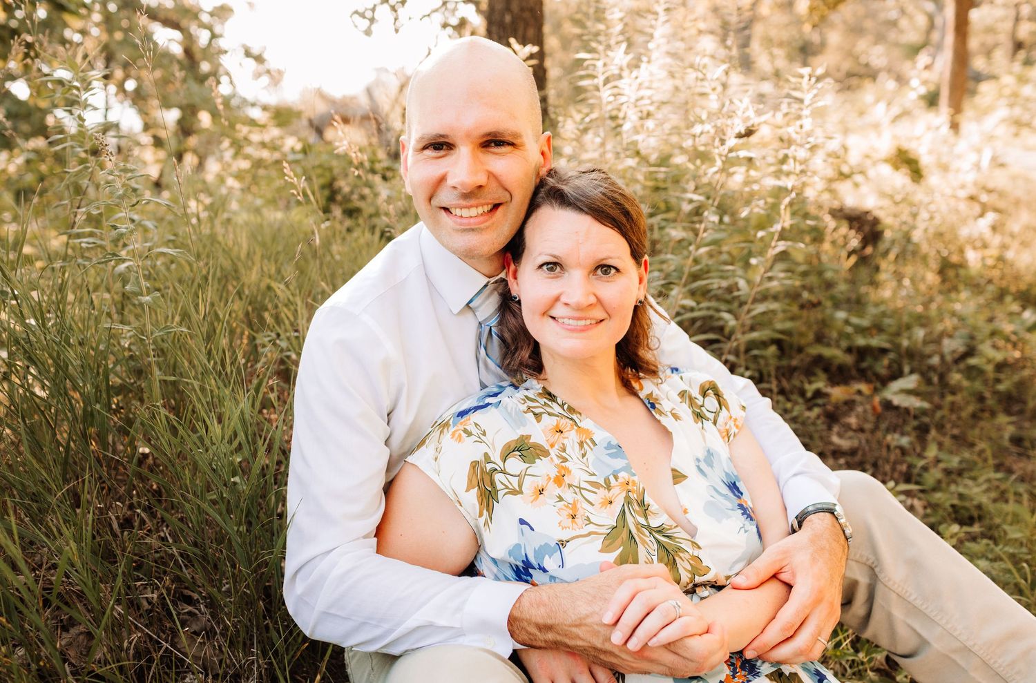 Smiling couple sitting together in a sunlit meadow, woman in floral dress and man in light blue shirt.