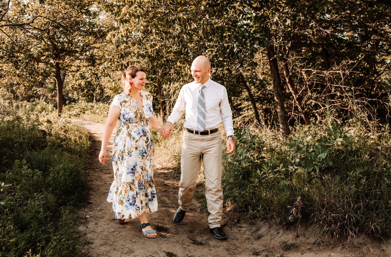 Happy couple holding hands and walking along a sunny forest path, woman in floral dress and man in khakis.
