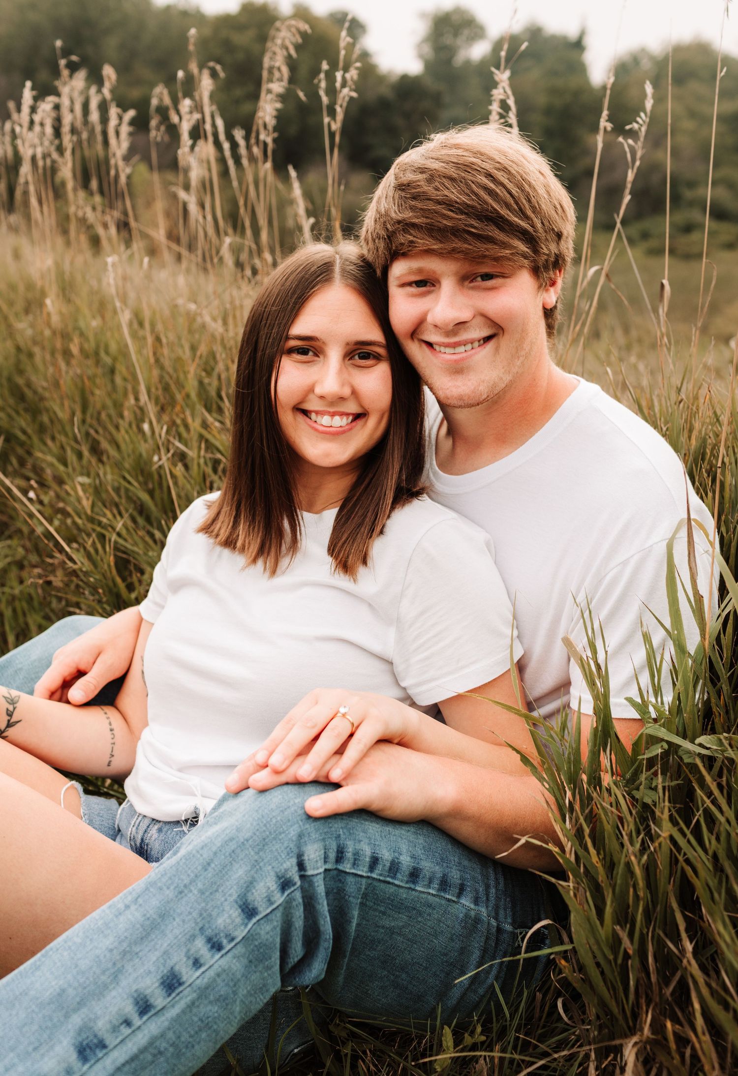 Happy young couple in white t-shirts sitting together in a tall grass field, smiling at camera.