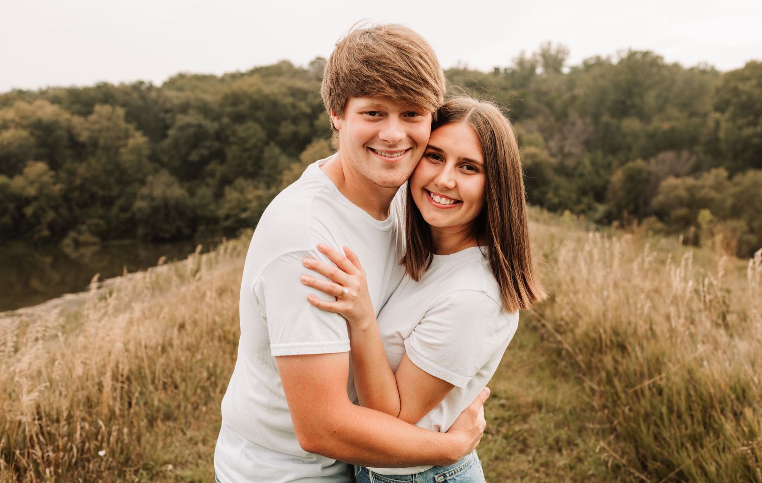 Young couple in white t-shirts embracing and smiling in a golden meadow with trees in background.