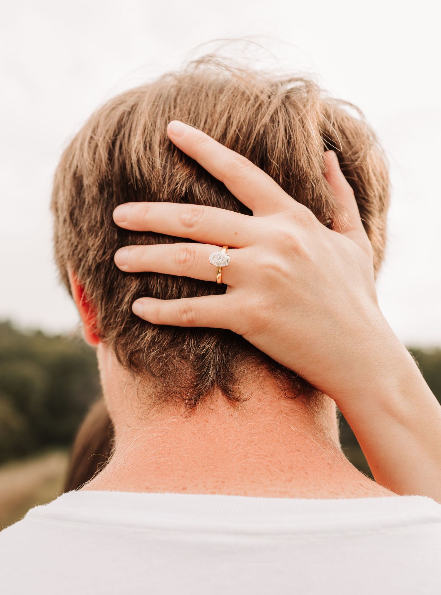 Close-up of a woman's hand with a diamond engagement ring resting on a man's head.