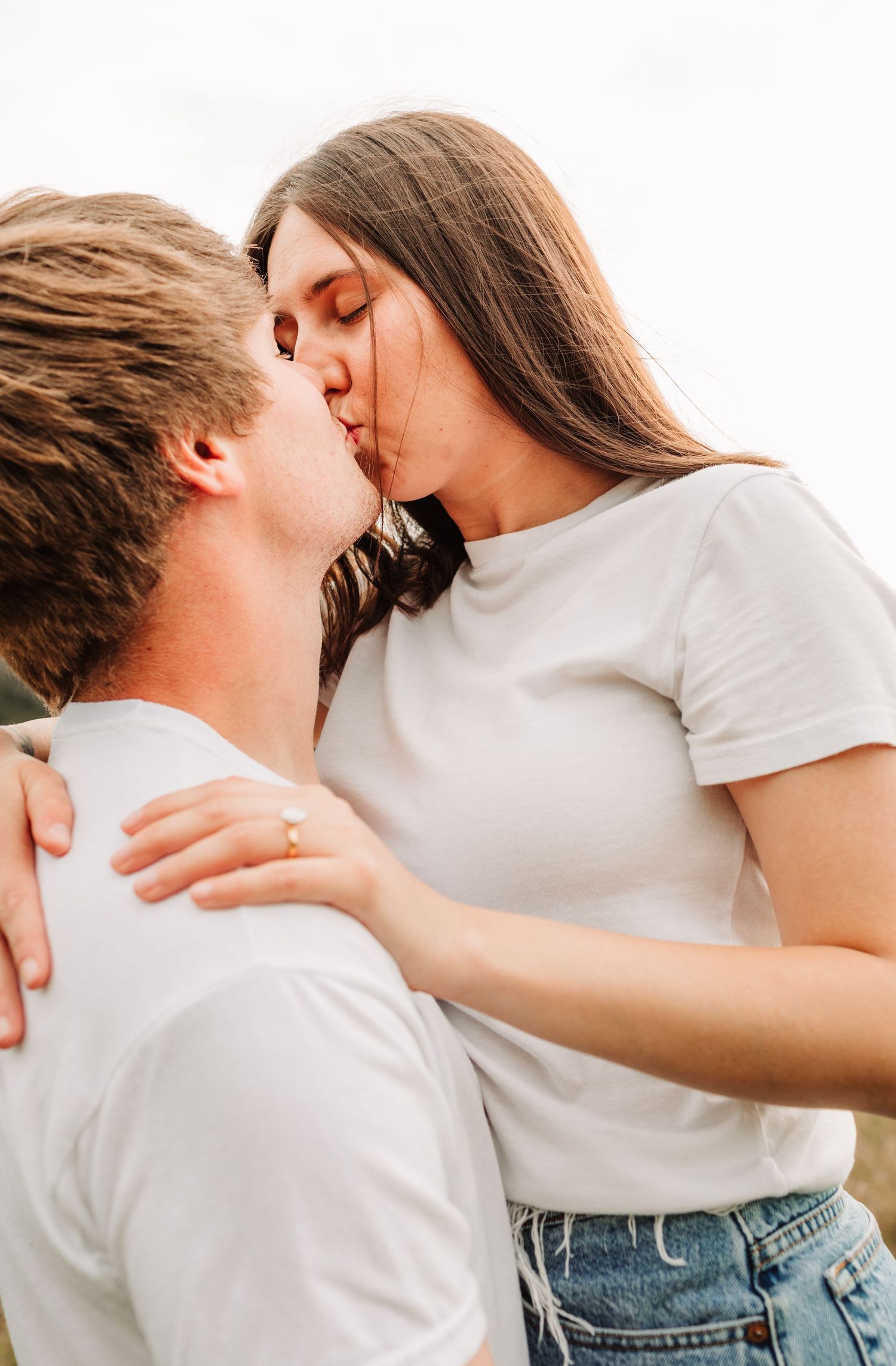 Young couple in white t-shirts sharing a romantic kiss outdoors, woman wearing engagement ring.