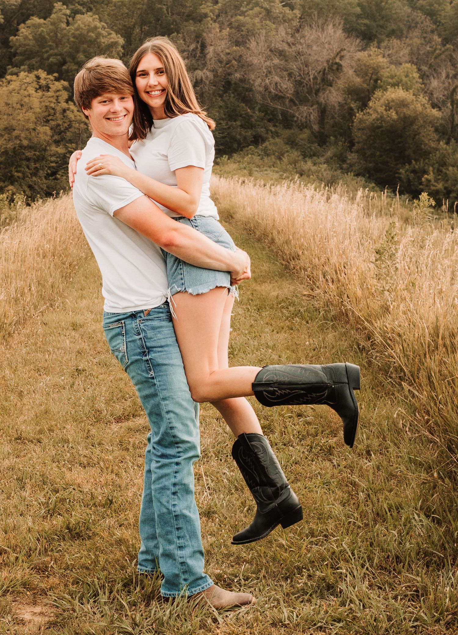 Young man lifting and hugging smiling woman in white t-shirt and cowboy boots in a golden field.