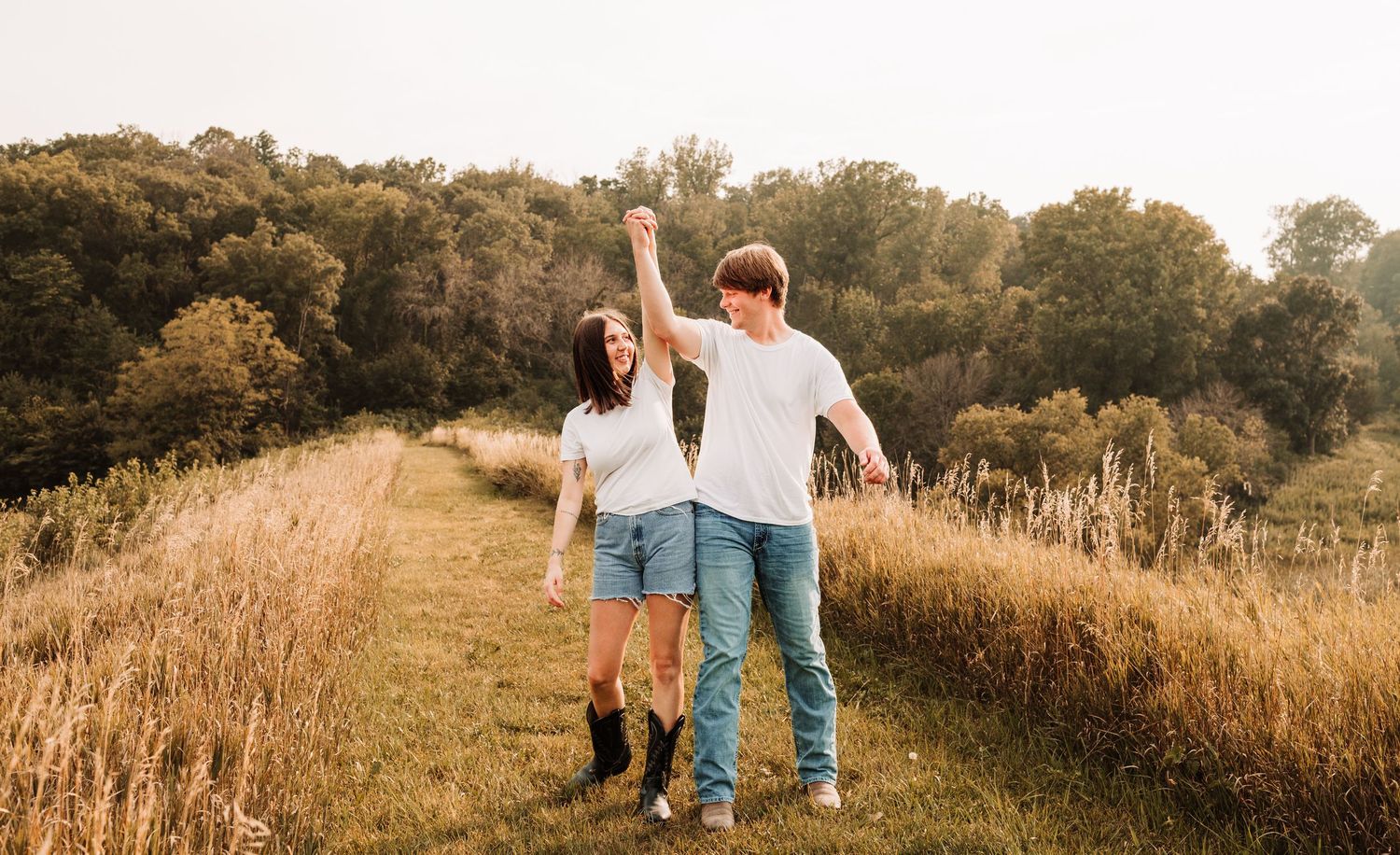 A joyful couple dances in a golden meadow, holding hands with arms raised, surrounded by lush green trees.