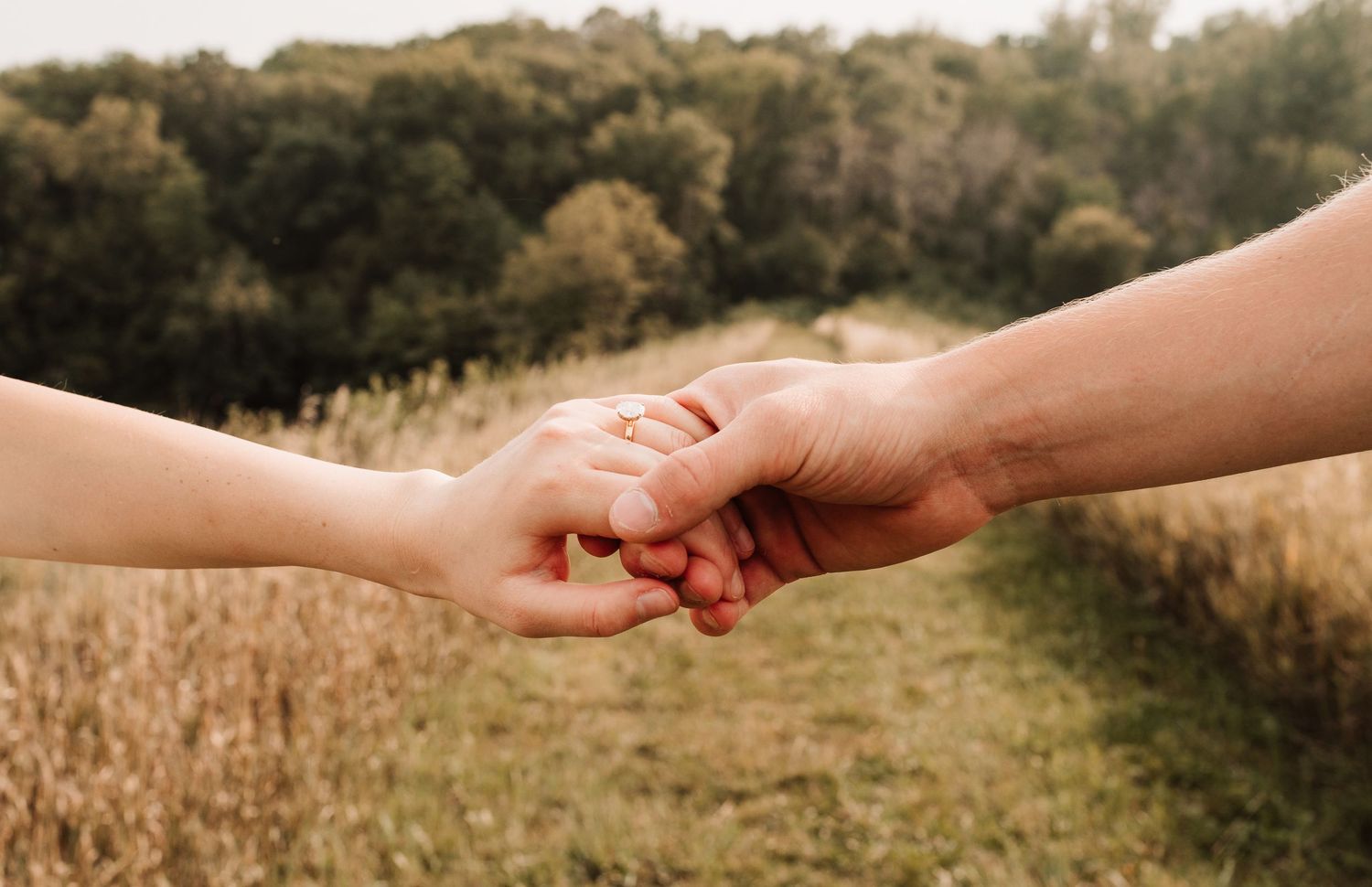 Close-up of a couple holding hands in a field, showcasing an engagement ring on the woman's finger.