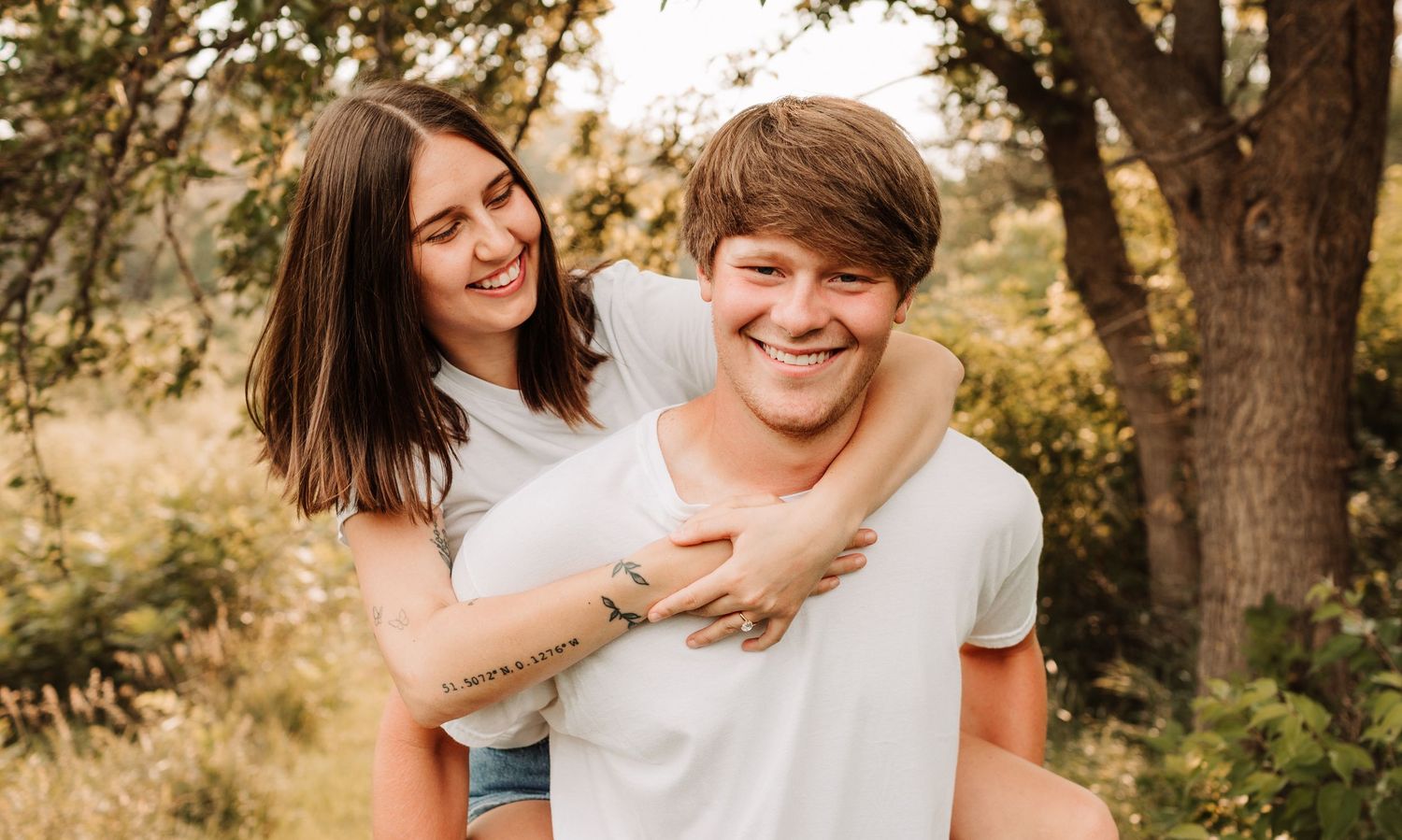 A smiling young woman hugs a laughing man from behind in a sunlit outdoor setting with trees.