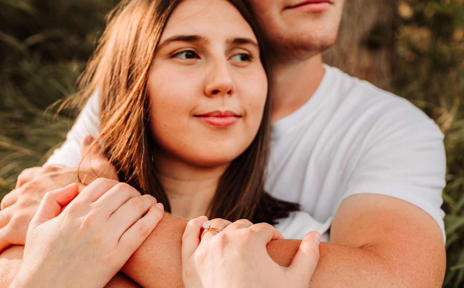 A couple embraces closely outdoors, the man wrapping his arms around the woman from behind in warm light.