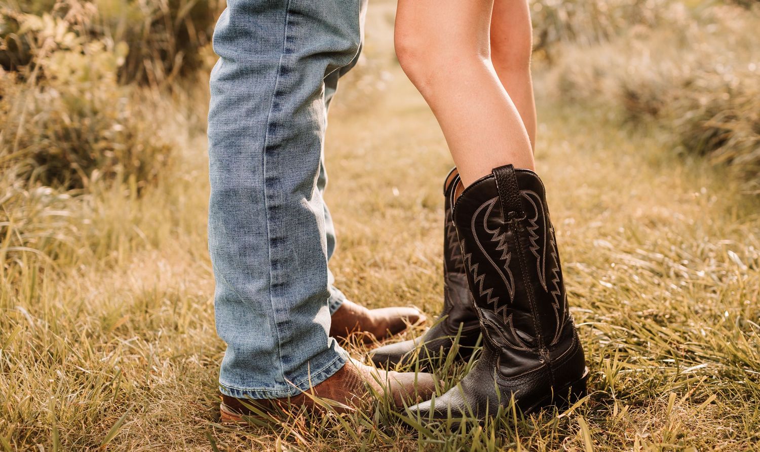 Close-up of a couple's feet, one wearing black cowboy boots and the other in jeans, standing in grass.