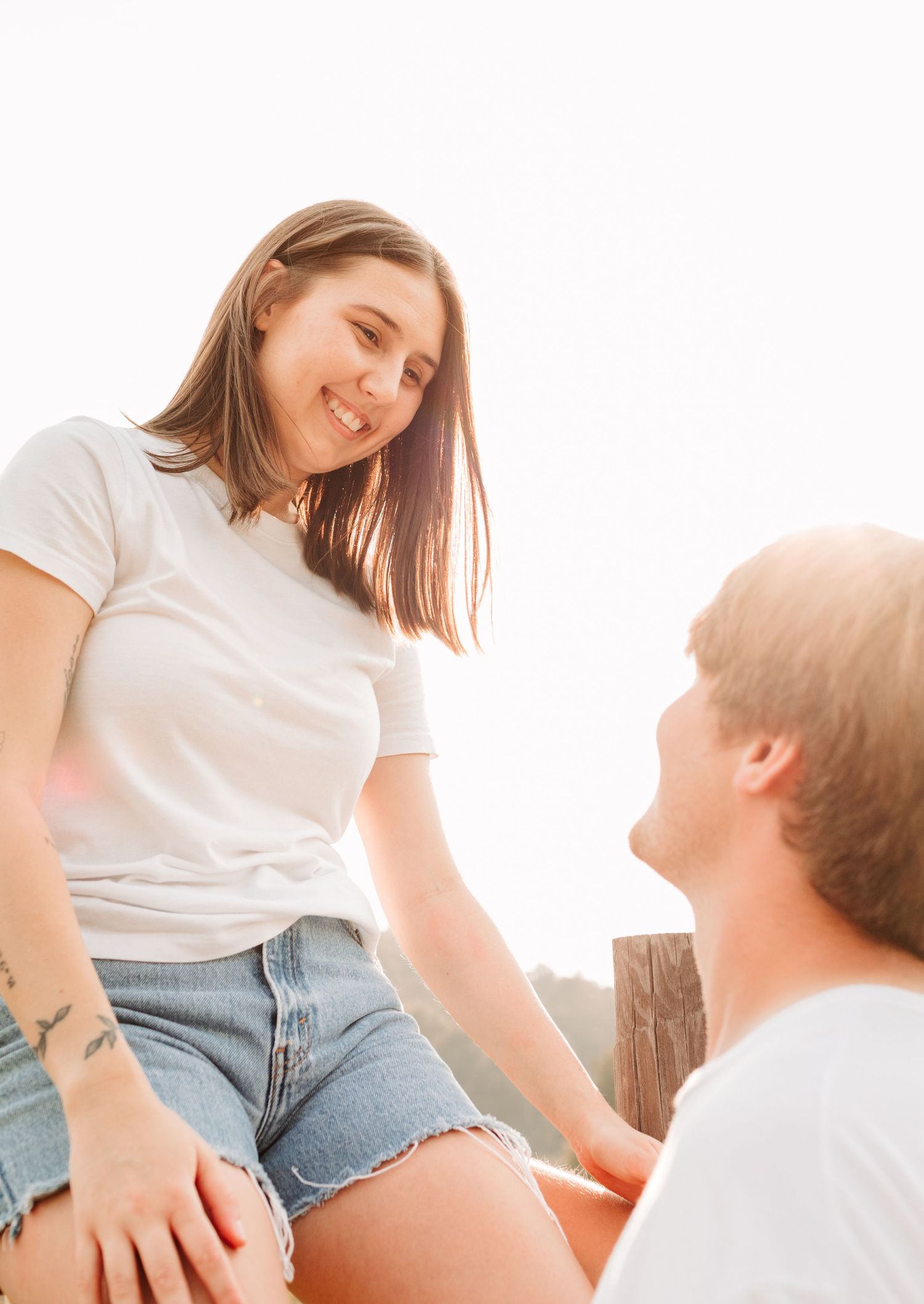 A smiling woman in a white tee and denim shorts sits on a fence, chatting with a man in bright sunlight.