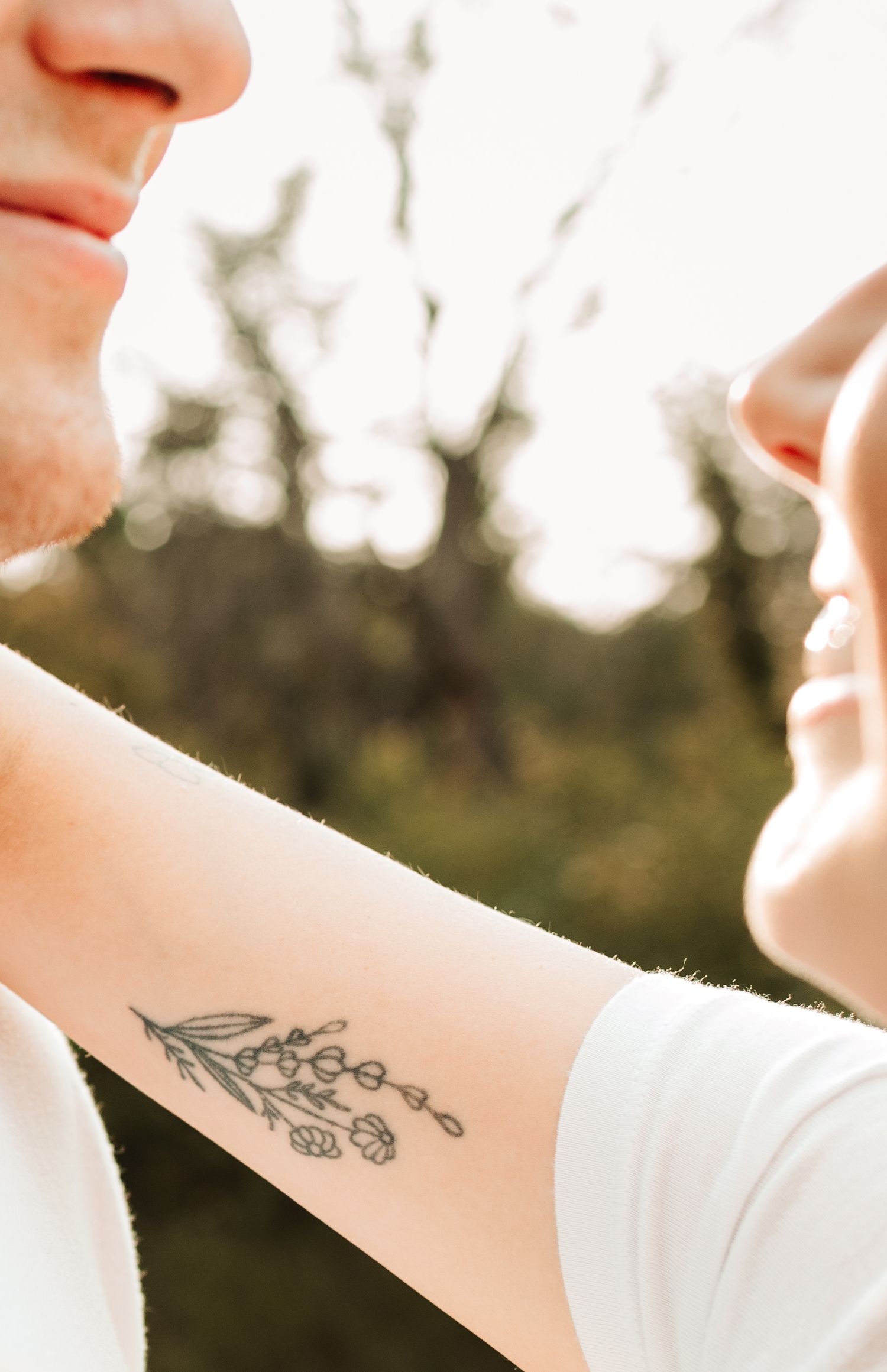 Close-up of a woman's forearm tattoo of flowers as a couple faces each other outdoors with blurred trees.