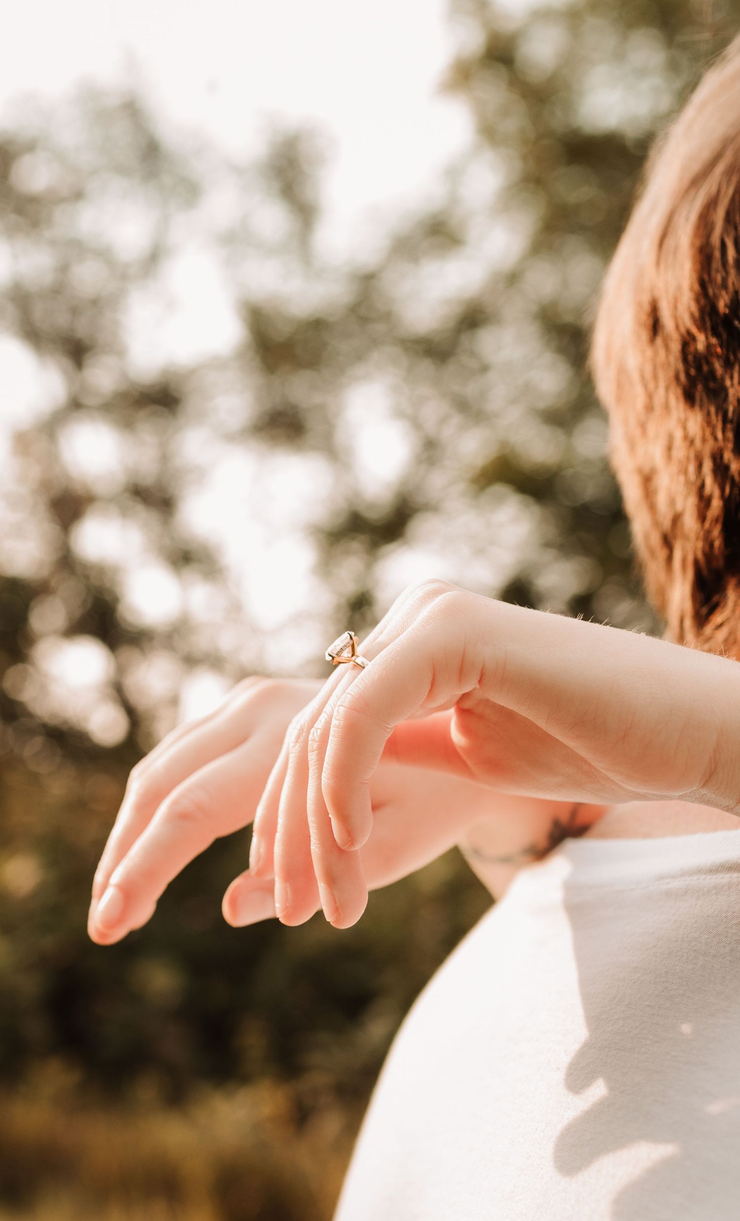 Close-up of a woman's hand displaying a delicate gold engagement ring outdoors in warm, soft lighting.