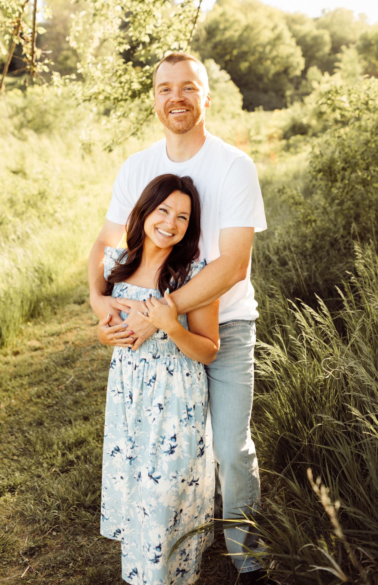 Smiling couple embracing outdoors in lush green field, woman in floral blue dress, man in white tee and jeans.