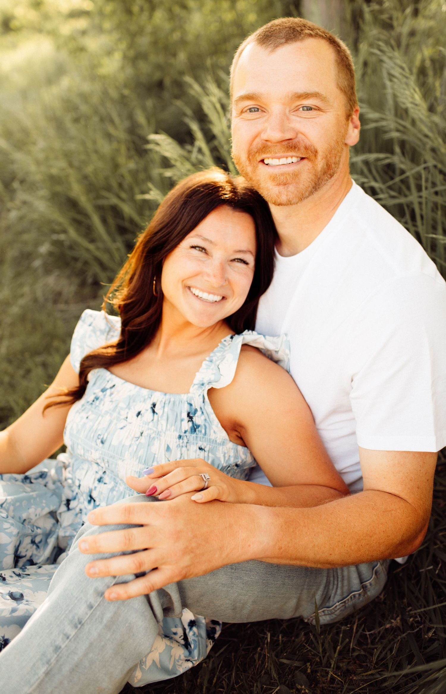 Happy couple sitting outdoors in tall green grass, woman in floral dress and man in white t-shirt smiling warmly.