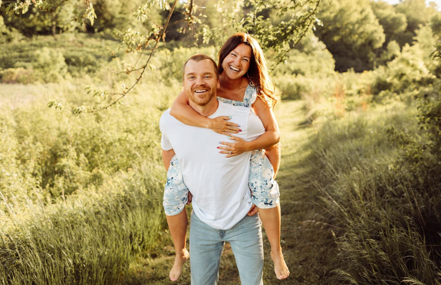 Happy couple laughing outdoors, man giving woman a piggyback ride through a sunny green meadow path.