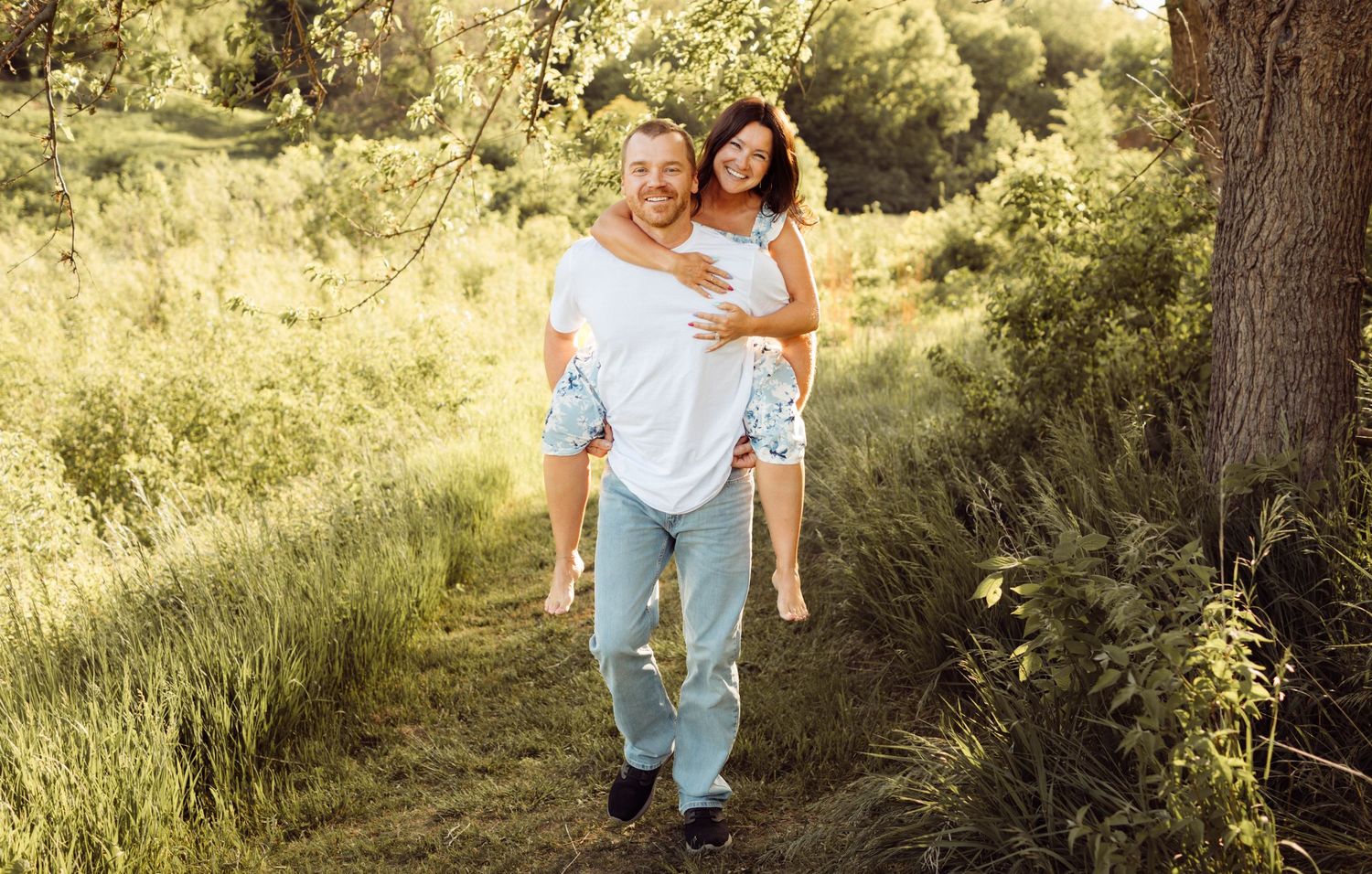 Joyful couple in nature, man carrying woman on his back along a sunlit green trail surrounded by foliage.