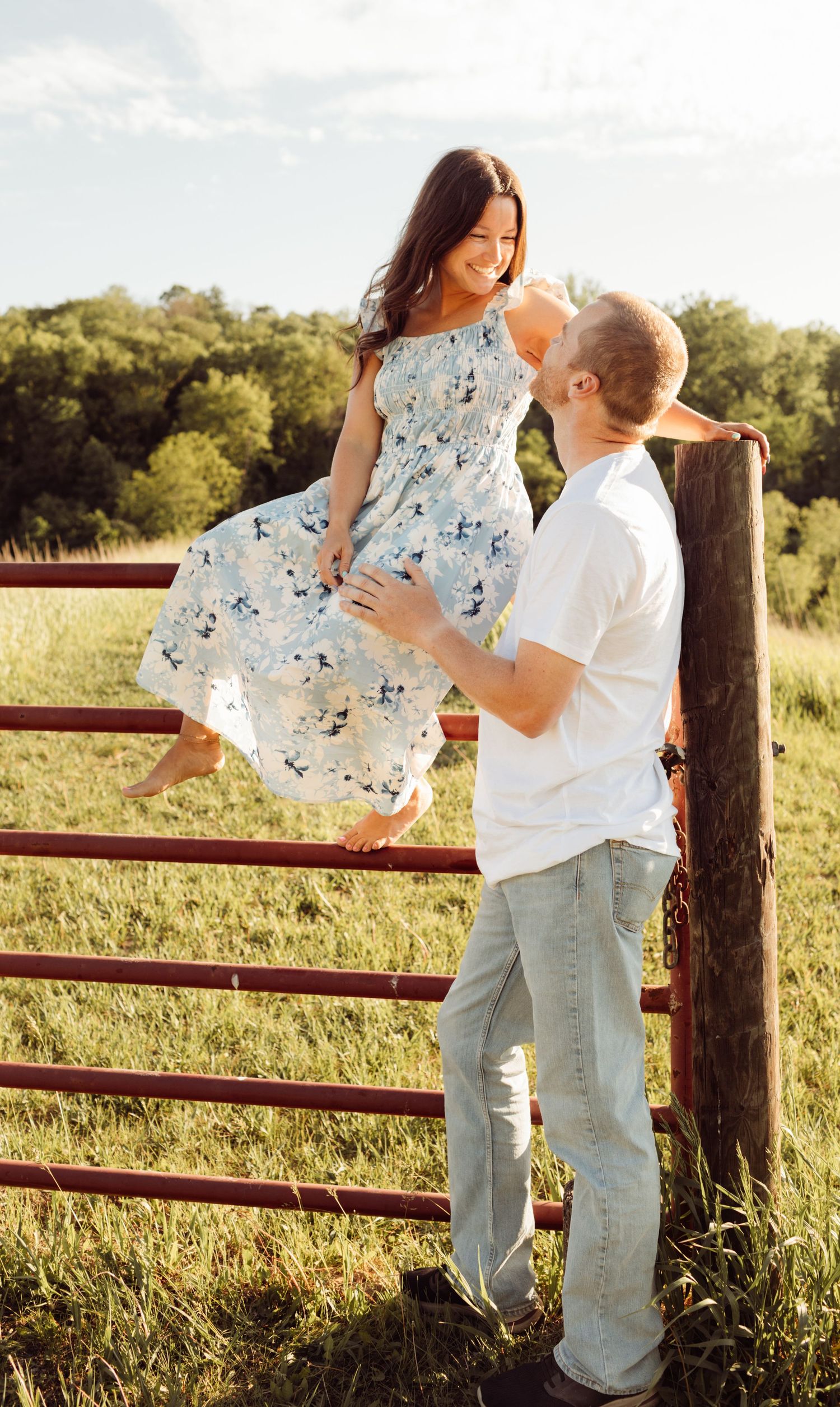 Woman in floral dress sitting on rustic fence, man in white shirt gazing up at her in sunny field.