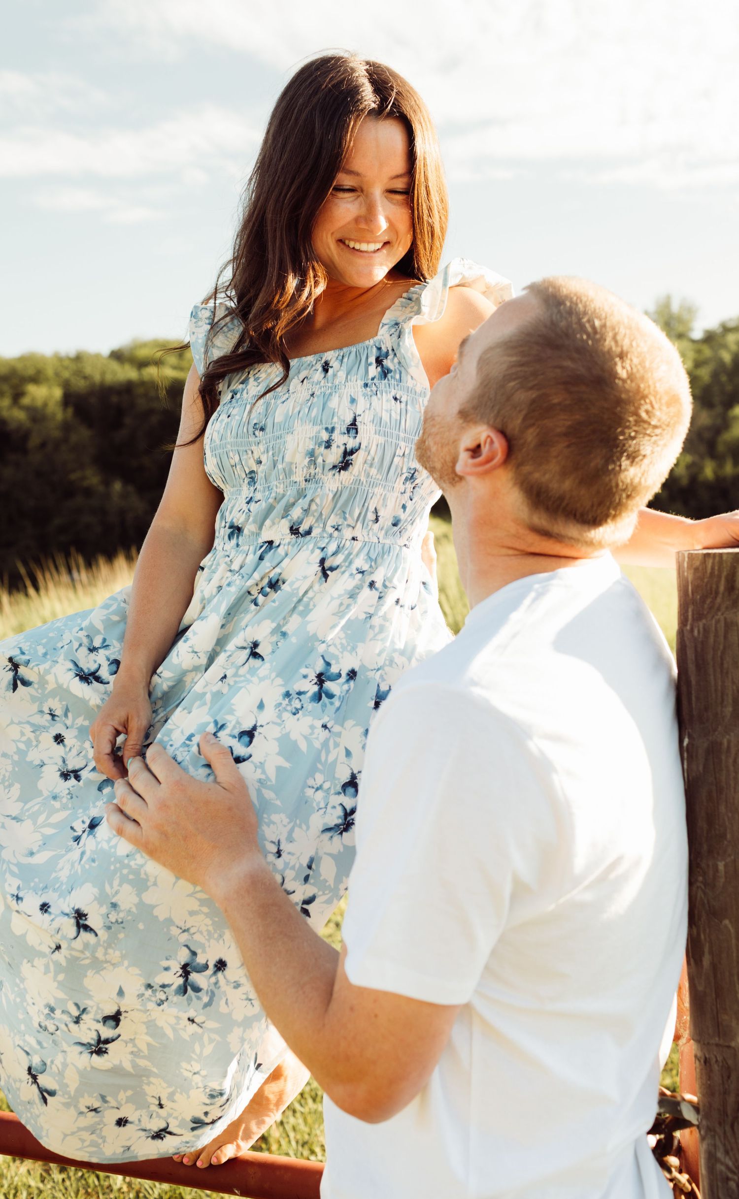 Couple sharing a tender moment by wooden fence, woman in blue floral dress smiling down at man outdoors.