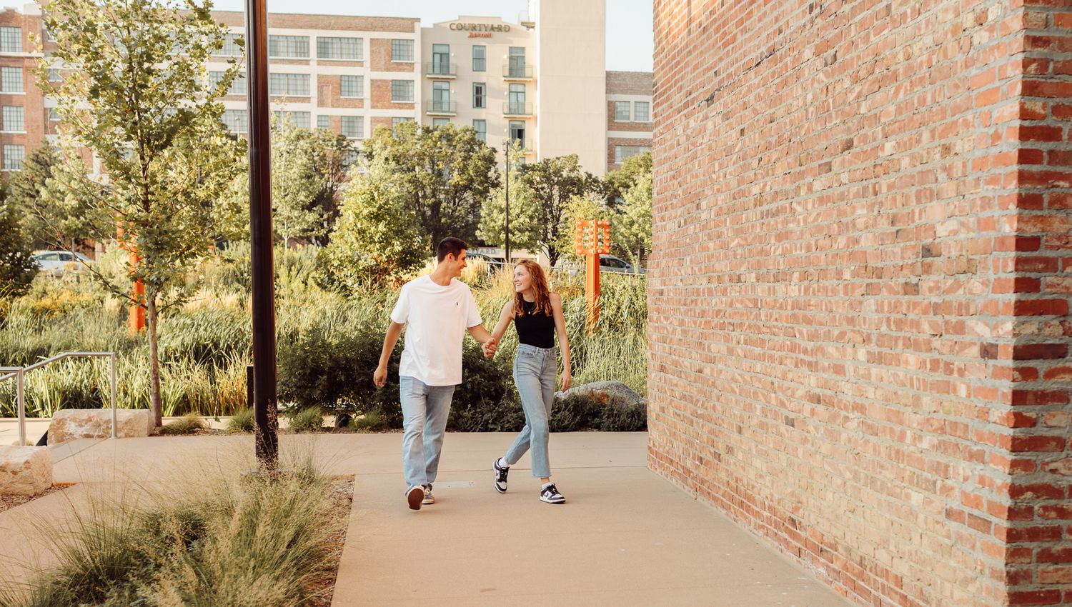 Young couple holding hands and walking along a sunny outdoor pathway beside a brick wall.