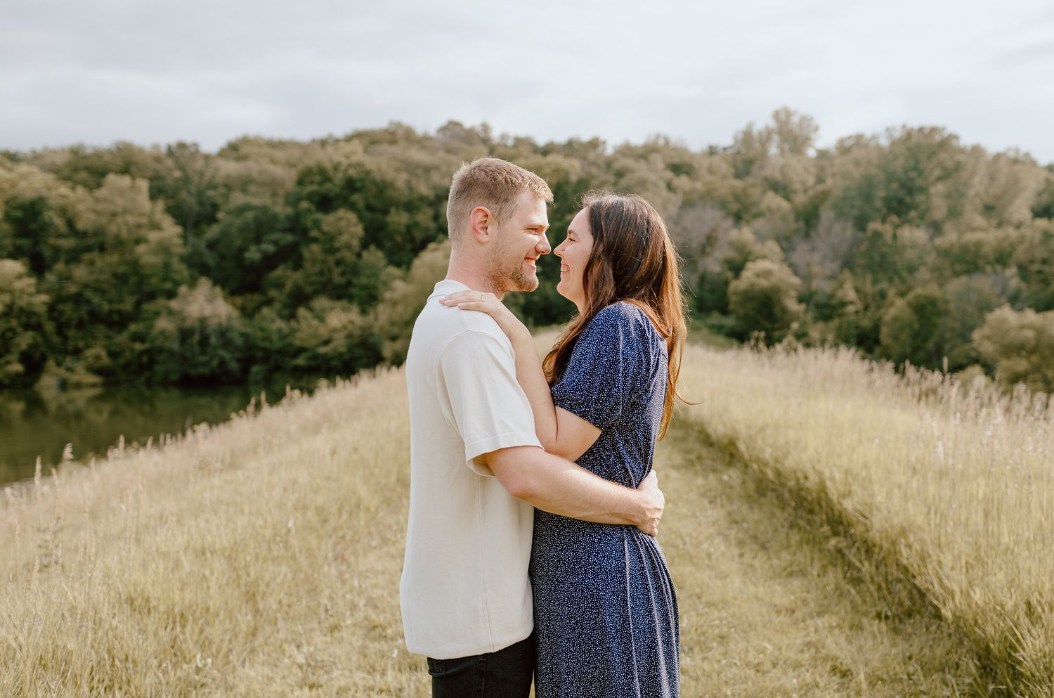 Young couple facing each other in a golden meadow near a river, sharing a romantic close moment.