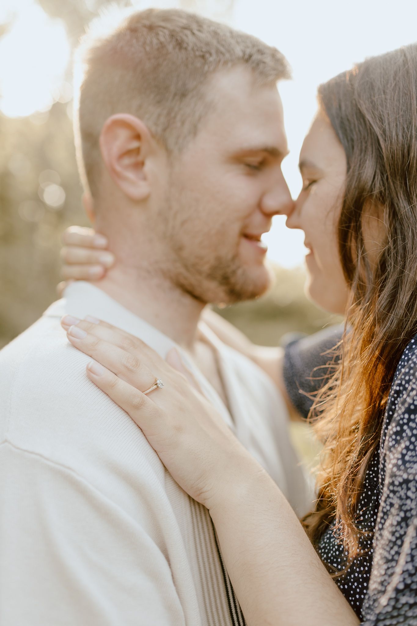 Engaged couple nearly kissing in golden sunlight, woman's engagement ring visible on man's shoulder.