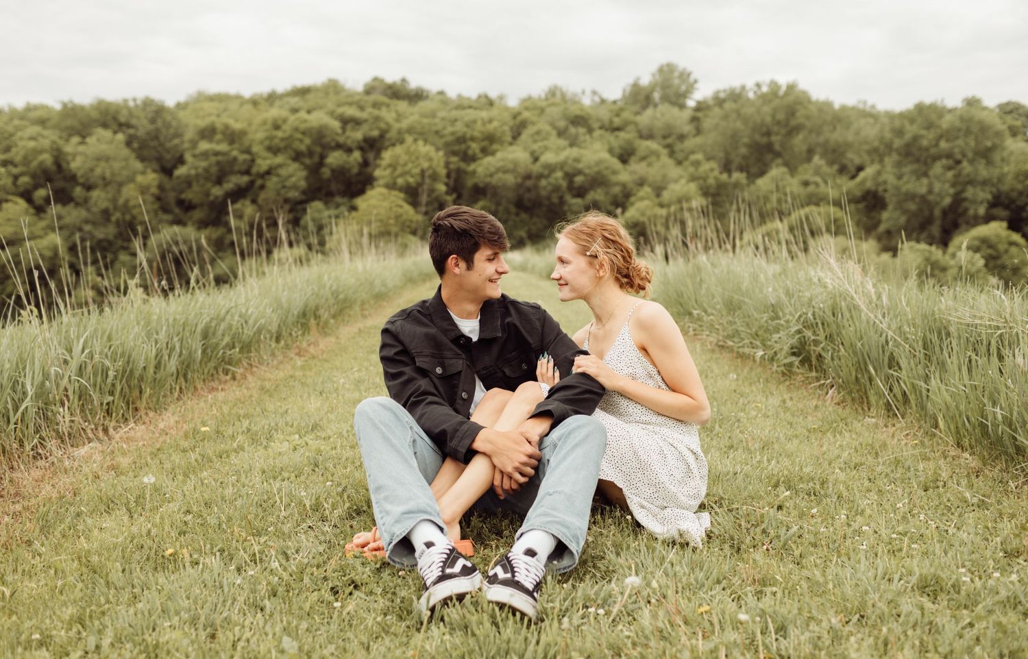 Young couple sitting face to face in grassy field path, smiling and sharing a romantic outdoor moment.