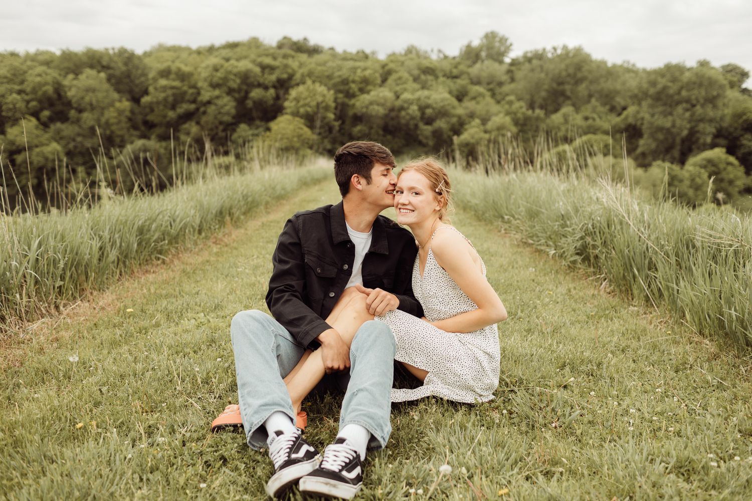 Young couple sitting close together in a green grass path, sharing a tender romantic outdoor moment.