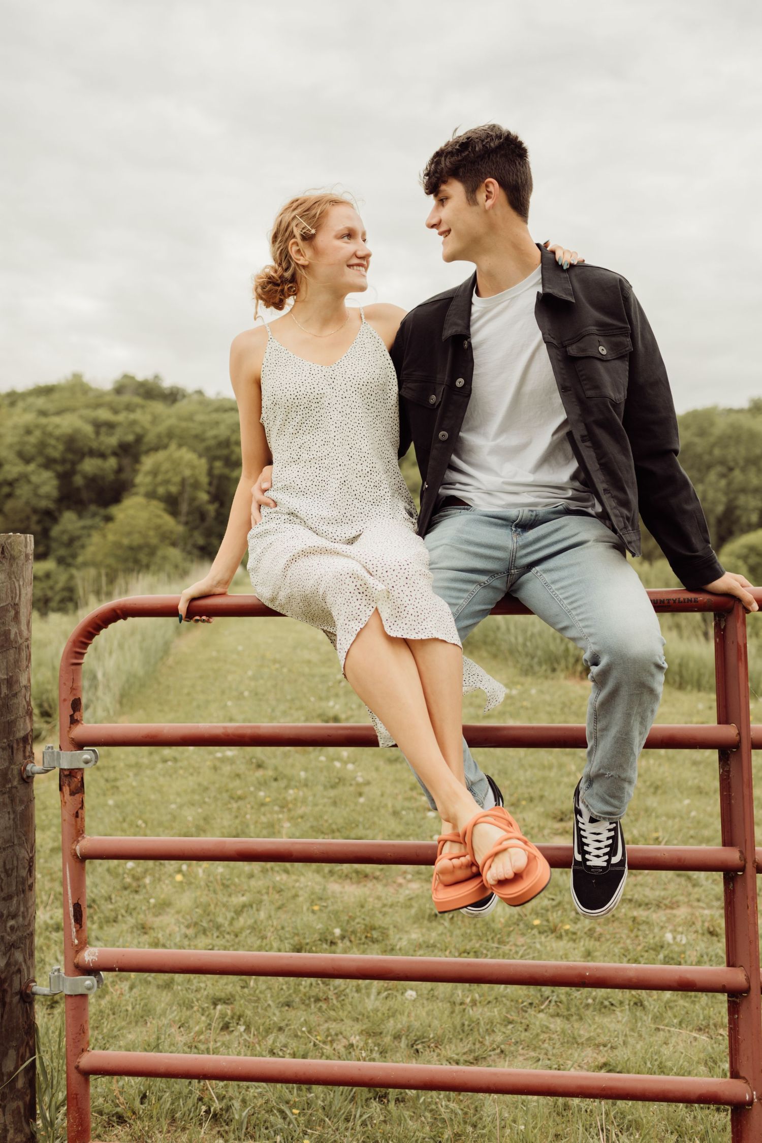 Young couple sitting on red metal farm gate, smiling at each other in an open green countryside field.