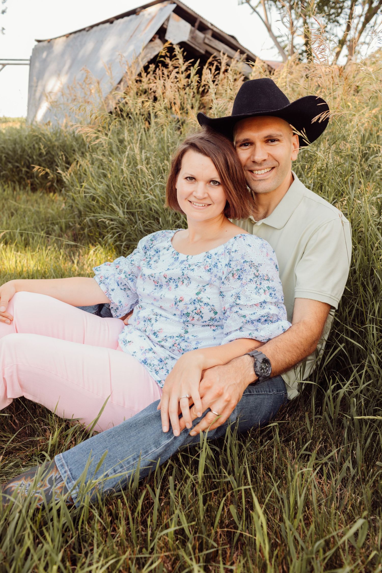 Smiling couple sitting in green grass near rustic barn, man in black cowboy hat embracing woman in floral top.